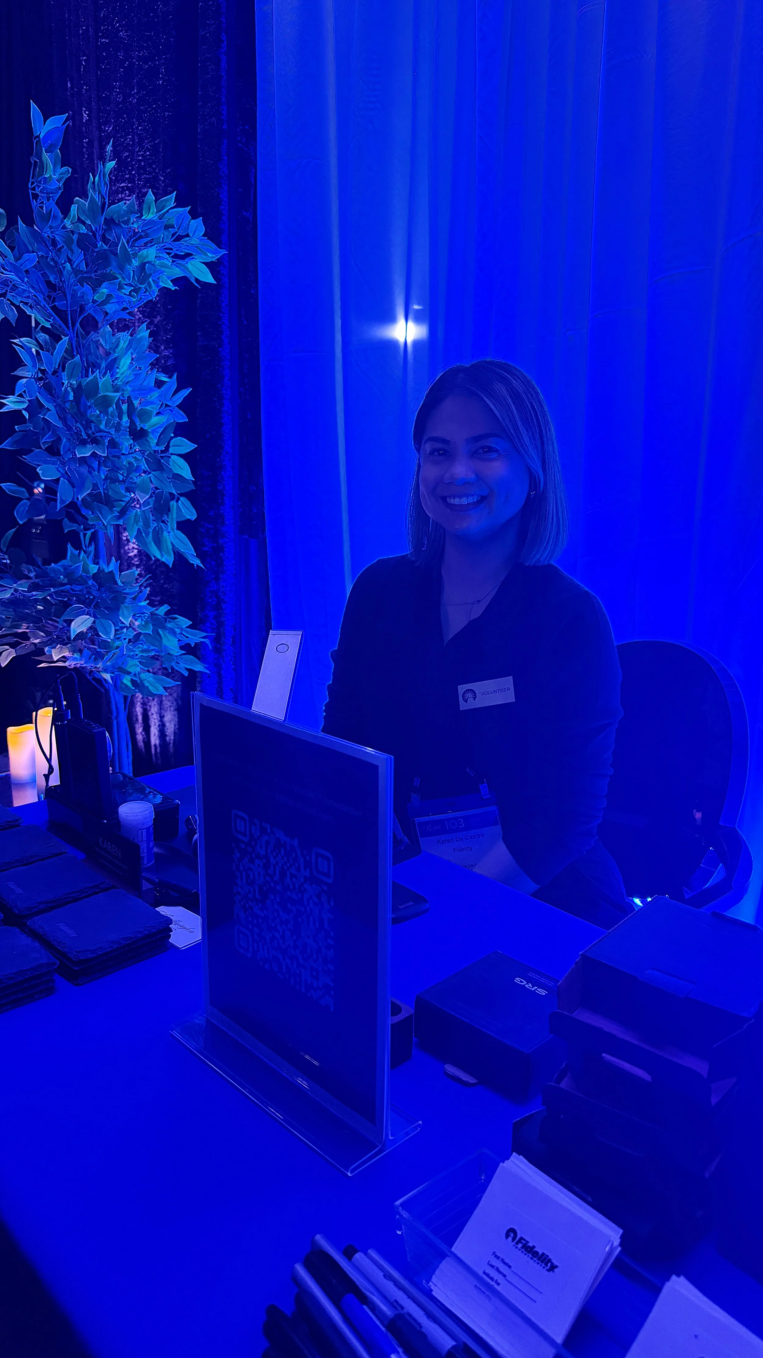 A woman sitting at a registration desk, smiling, with a tented background and blue lighting, wearing a volunteer badge.