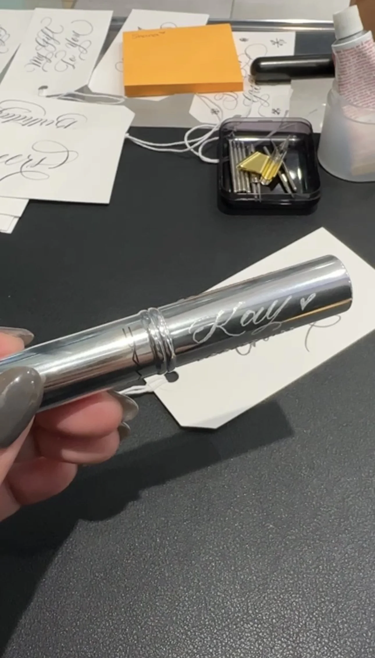 A metallic cylindrical object with the name 'Kay' engraved on it, held in a person's hand with gray painted nails. The background includes sheets of paper with calligraphy, a yellow sticky note, a black container with calligraphy supplies, and a whit