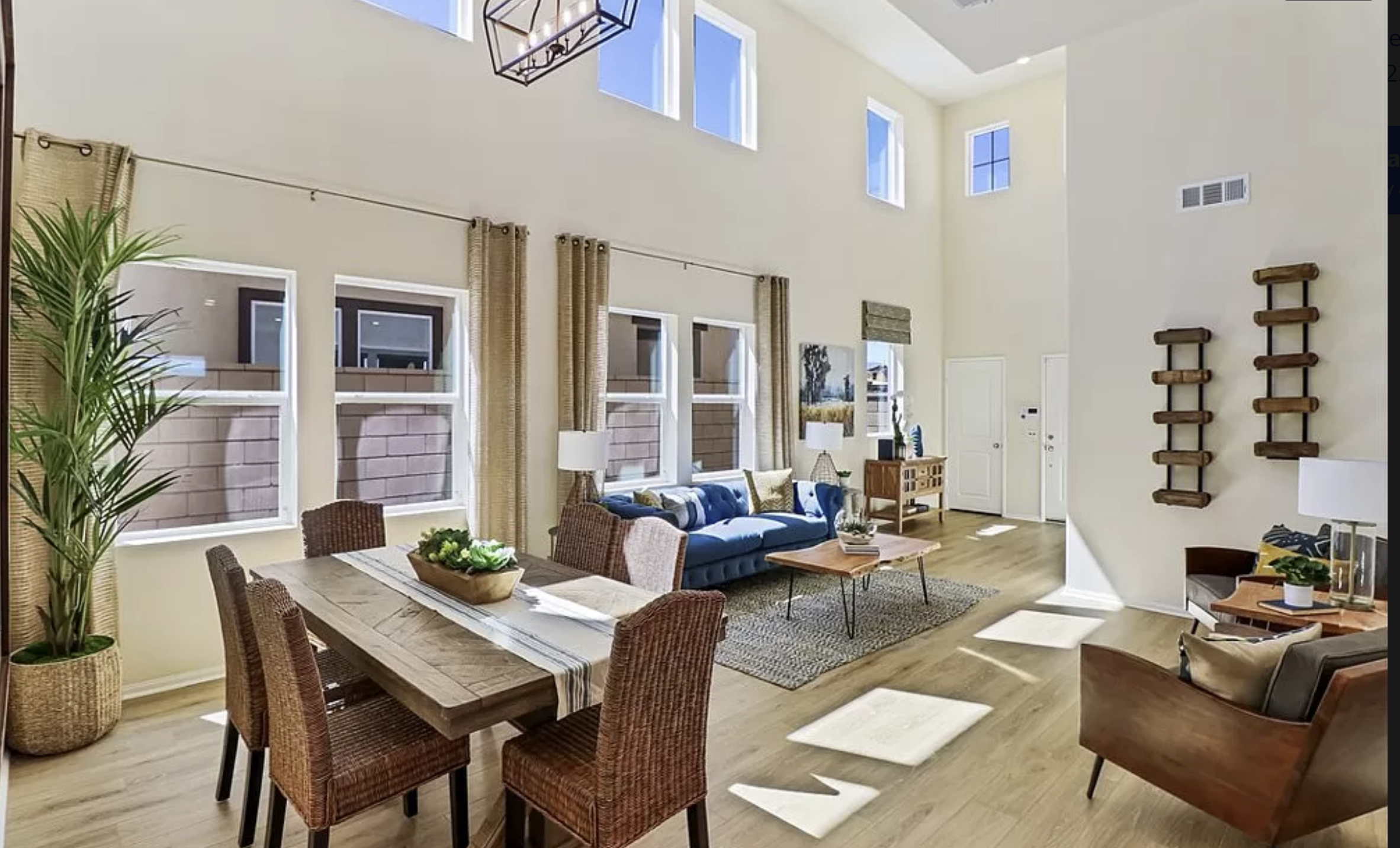 Bright living room with high ceilings, multiple windows, and modern decor including a blue sofa, wicker chairs, and indoor plants.
