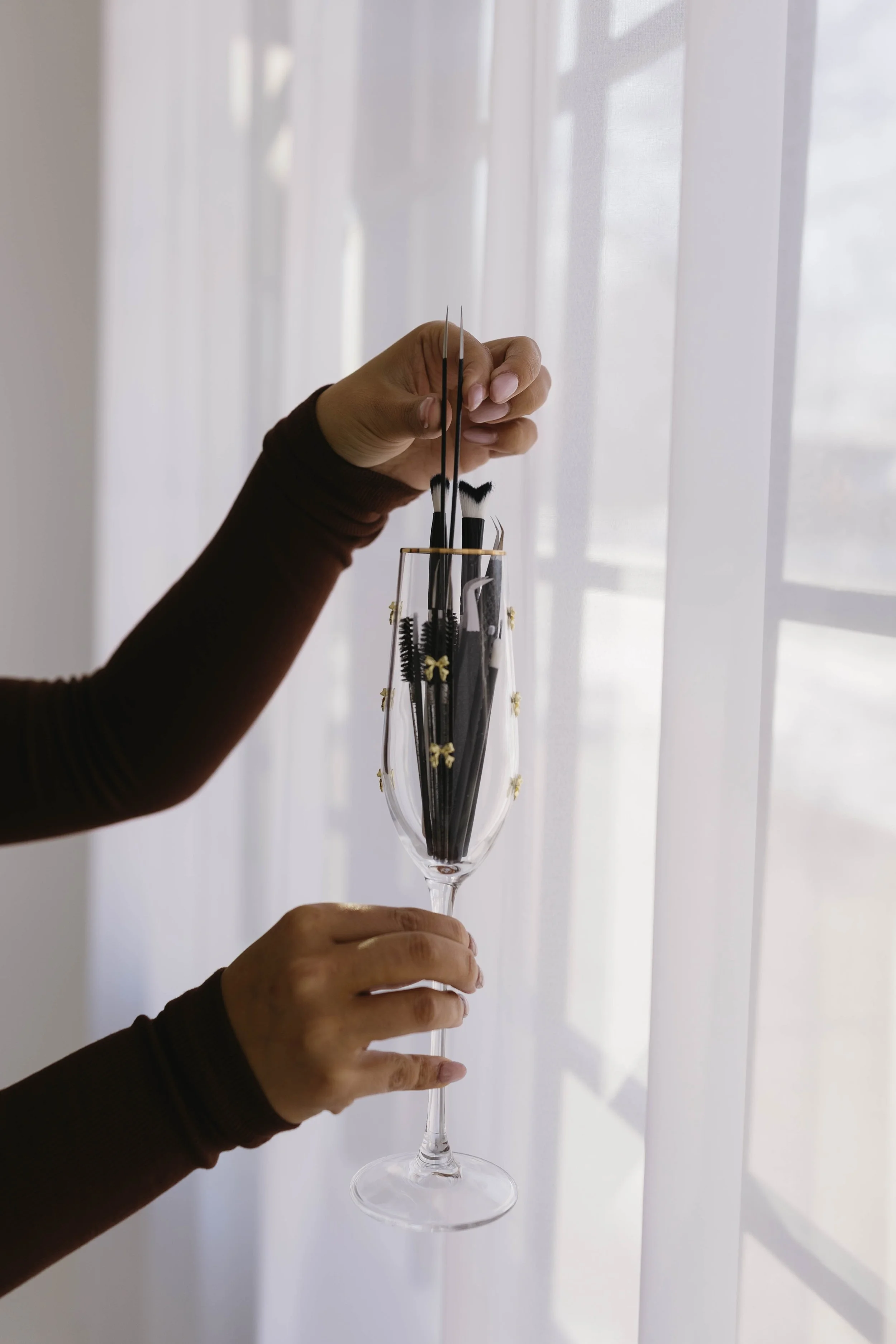 Person holding a champagne flute filled with makeup brushes in front of sheer curtains.