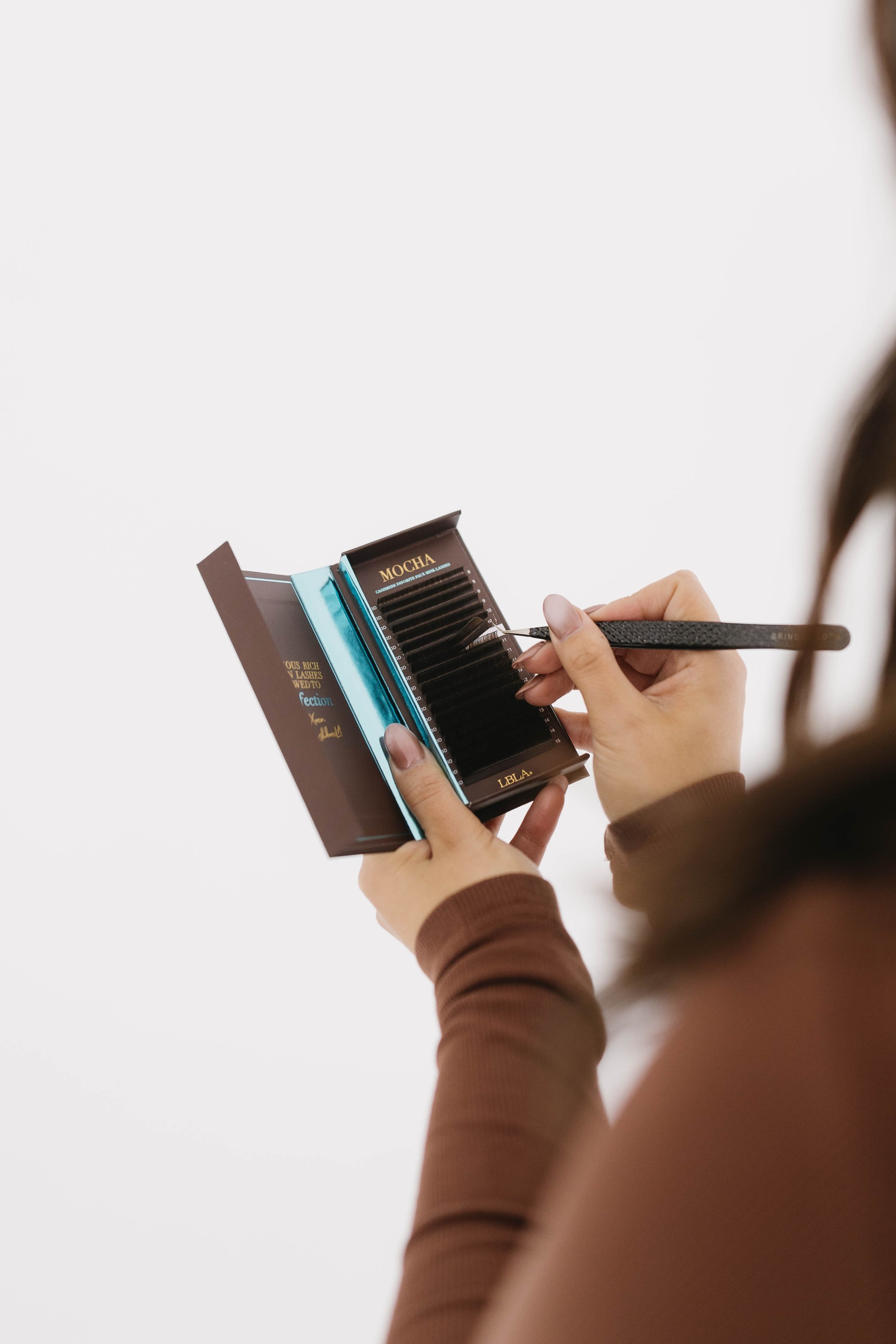 A person holds a small black box with several eyelash extensions and uses tweezers to pick one.