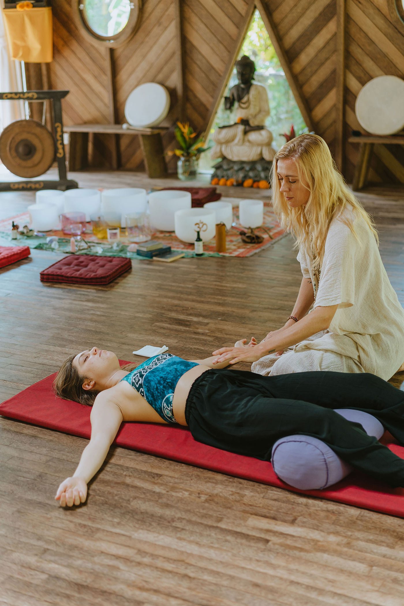 Woman practicing Reiki therapy on a person lying on a red mat in a meditation room with wooden walls, crystal bowls, a gong, and a statue in the background.