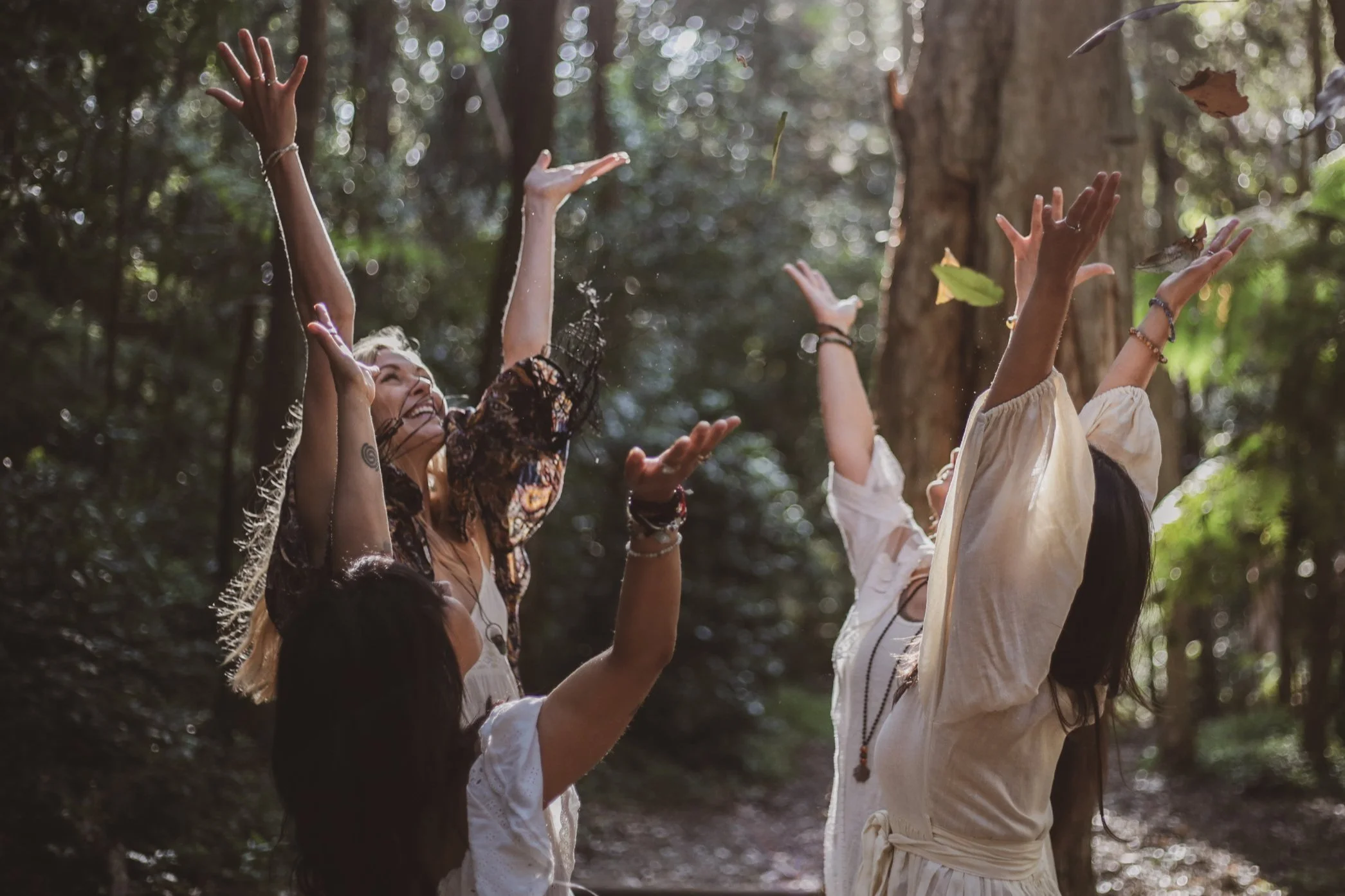 Group of women joyfully throwing leaves in a forest, wearing bohemian-style clothing, surrounded by lush greenery.