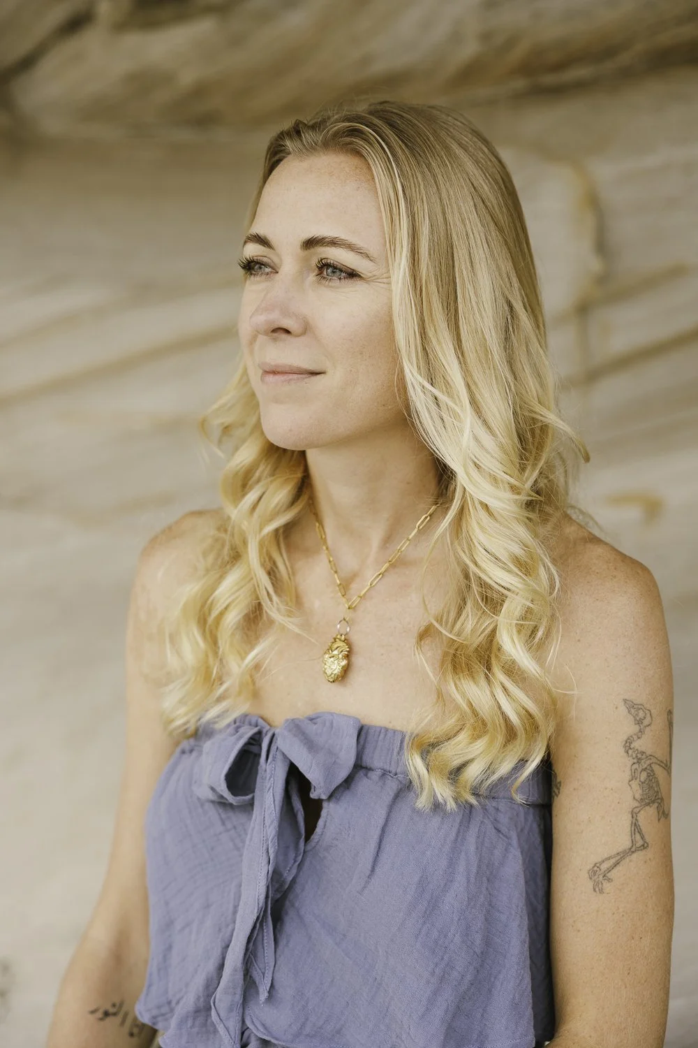 A woman with blonde, wavy hair gazing to the left, wearing a blue strapless top with a bow tied at the front, a gold necklace with a decorative pendant, and a tattoo of a skeleton on her left upper arm, in front of a wooden background.