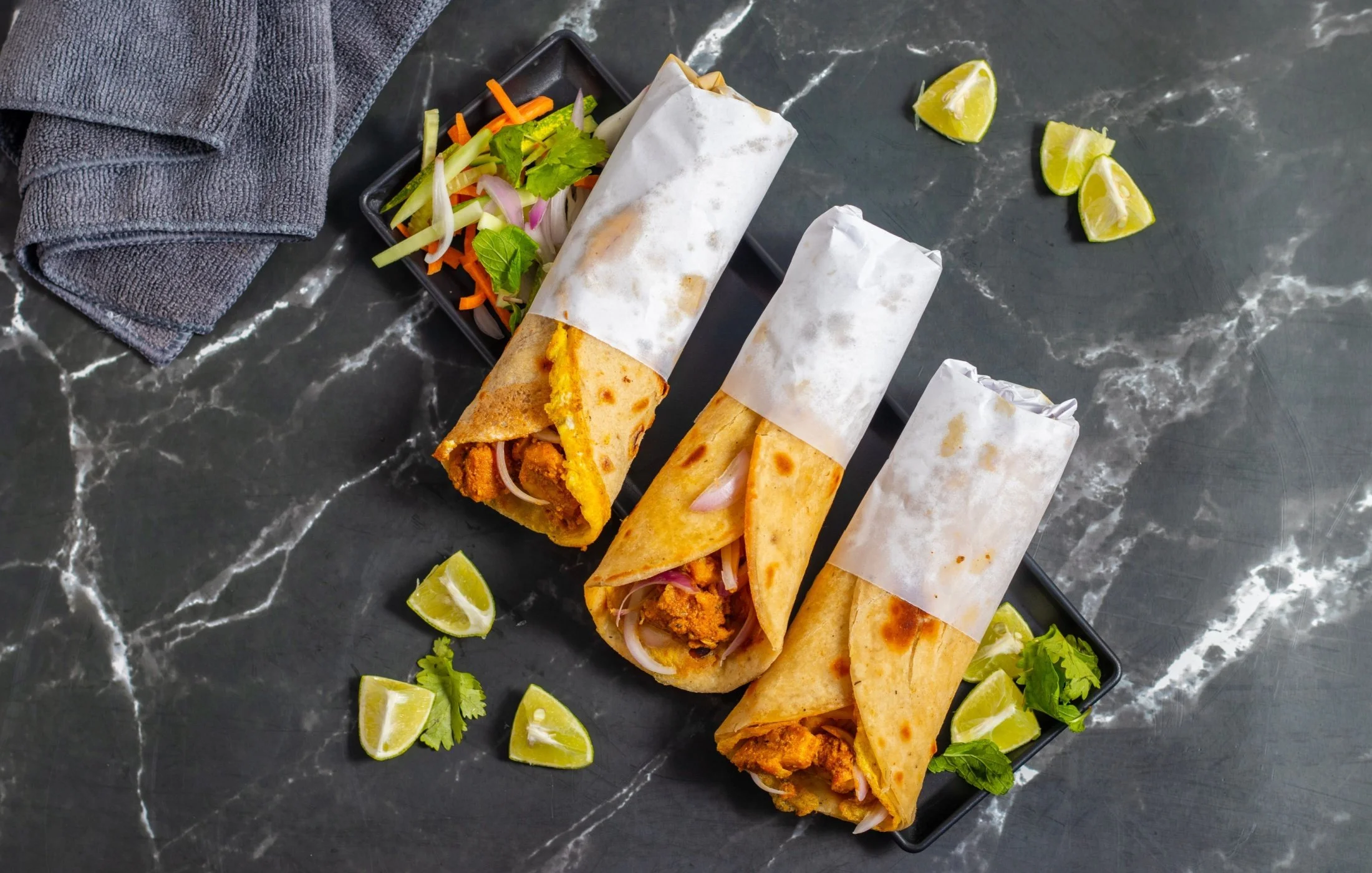 Overhead view of three freshly made chicken kathi rolls served with fresh salad, lime wedges, and herbs on a black platter, perfect for FIFA 2026 match-day dining.