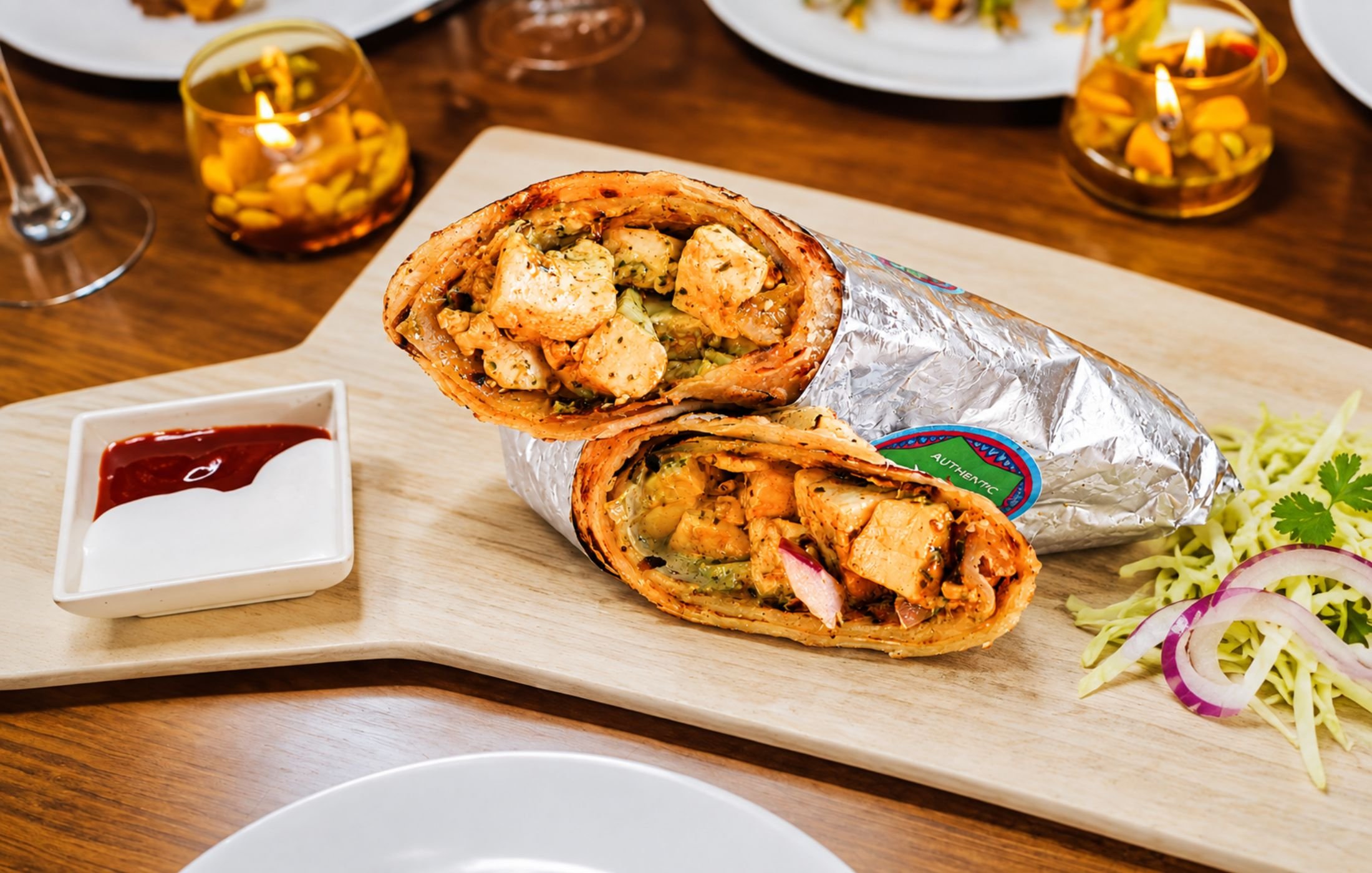 Close-up of a grilled chicken wrap filled with seasoned chicken, vegetables, and sauce, served on a wooden board with fresh shredded lettuce, red onion, and dipping sauce.