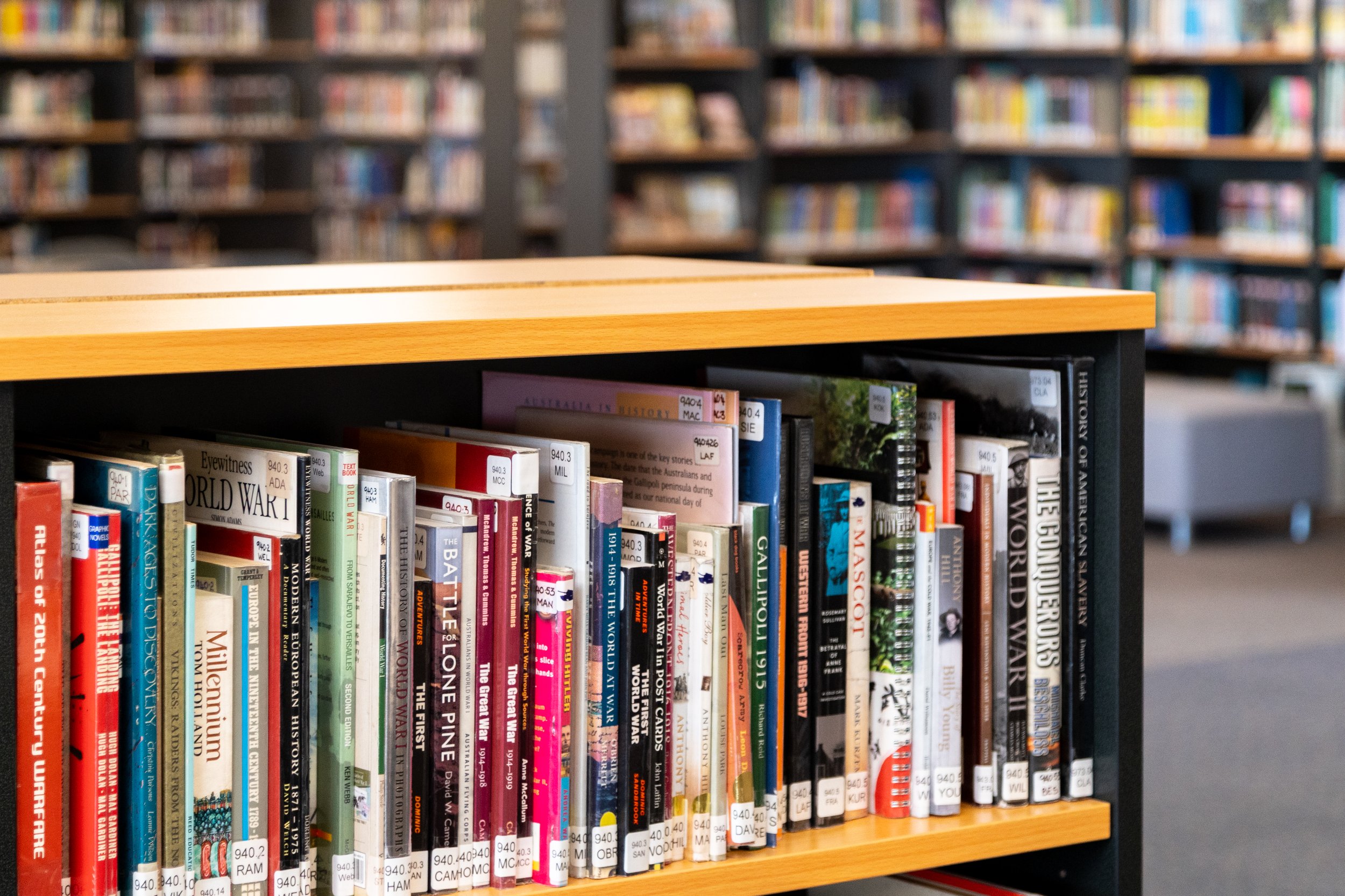 Bookshelves filled with books in a library, with a focus on a self-checkout station with a laptop and shopping cart in the background.