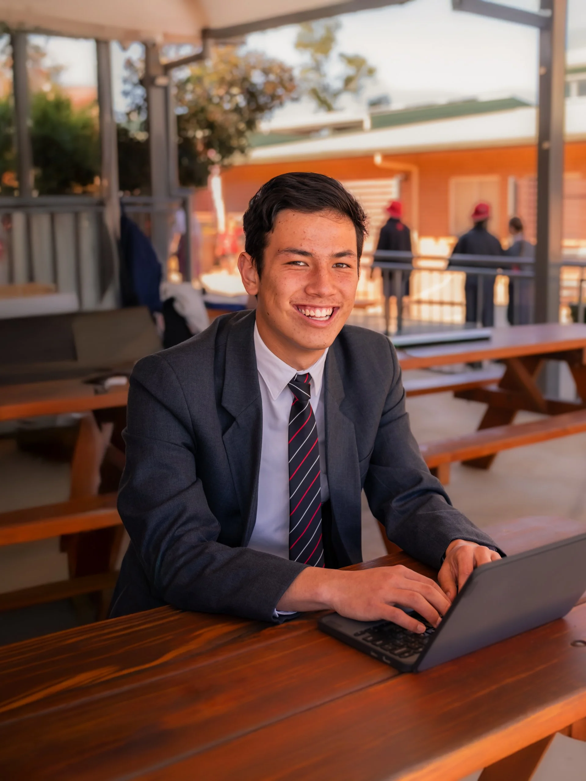 Young man in a suit sitting at a wooden table outdoors, working on a laptop and smiling.