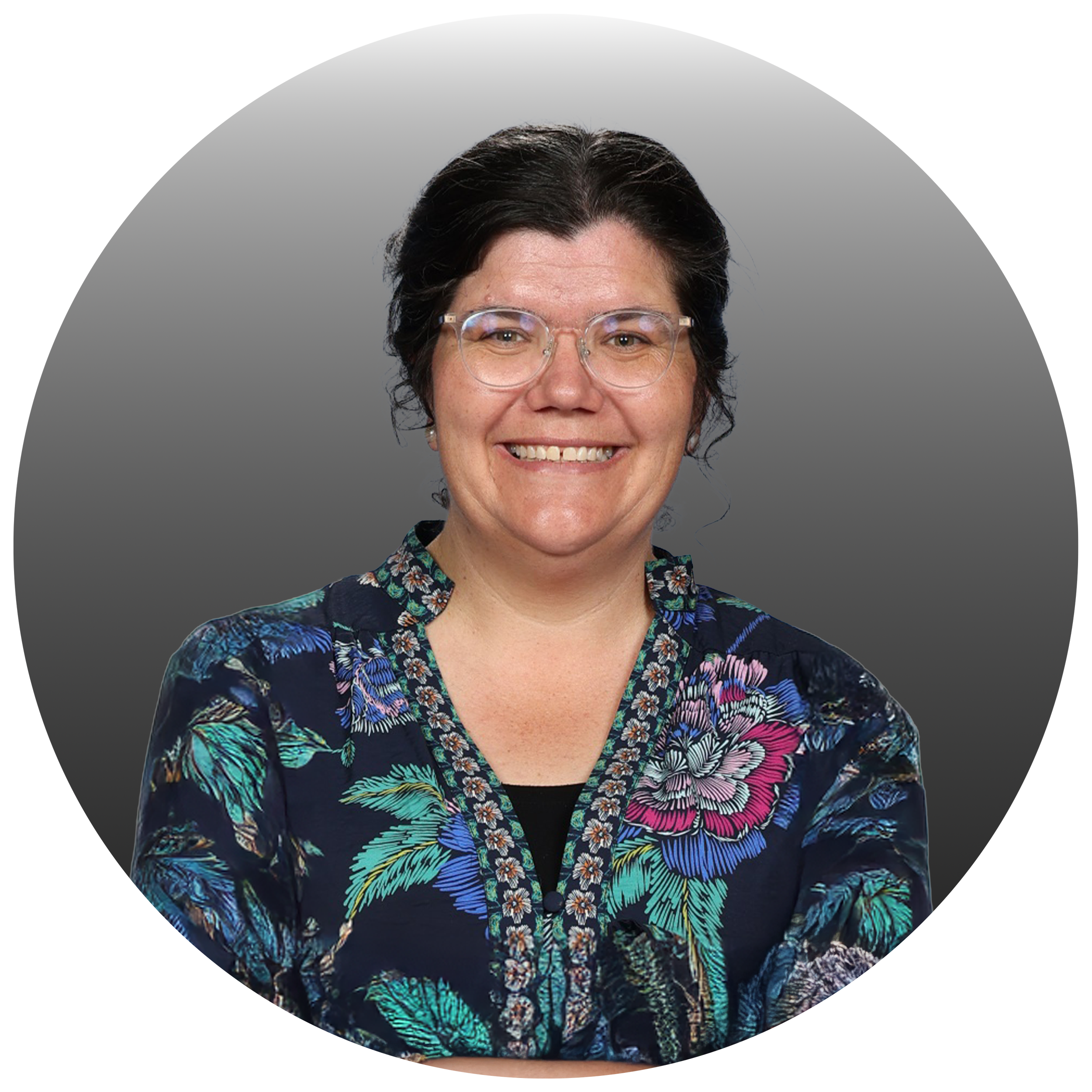 A woman with short dark hair, wearing glasses and a dark floral jacket, smiling for the camera against a gray background.