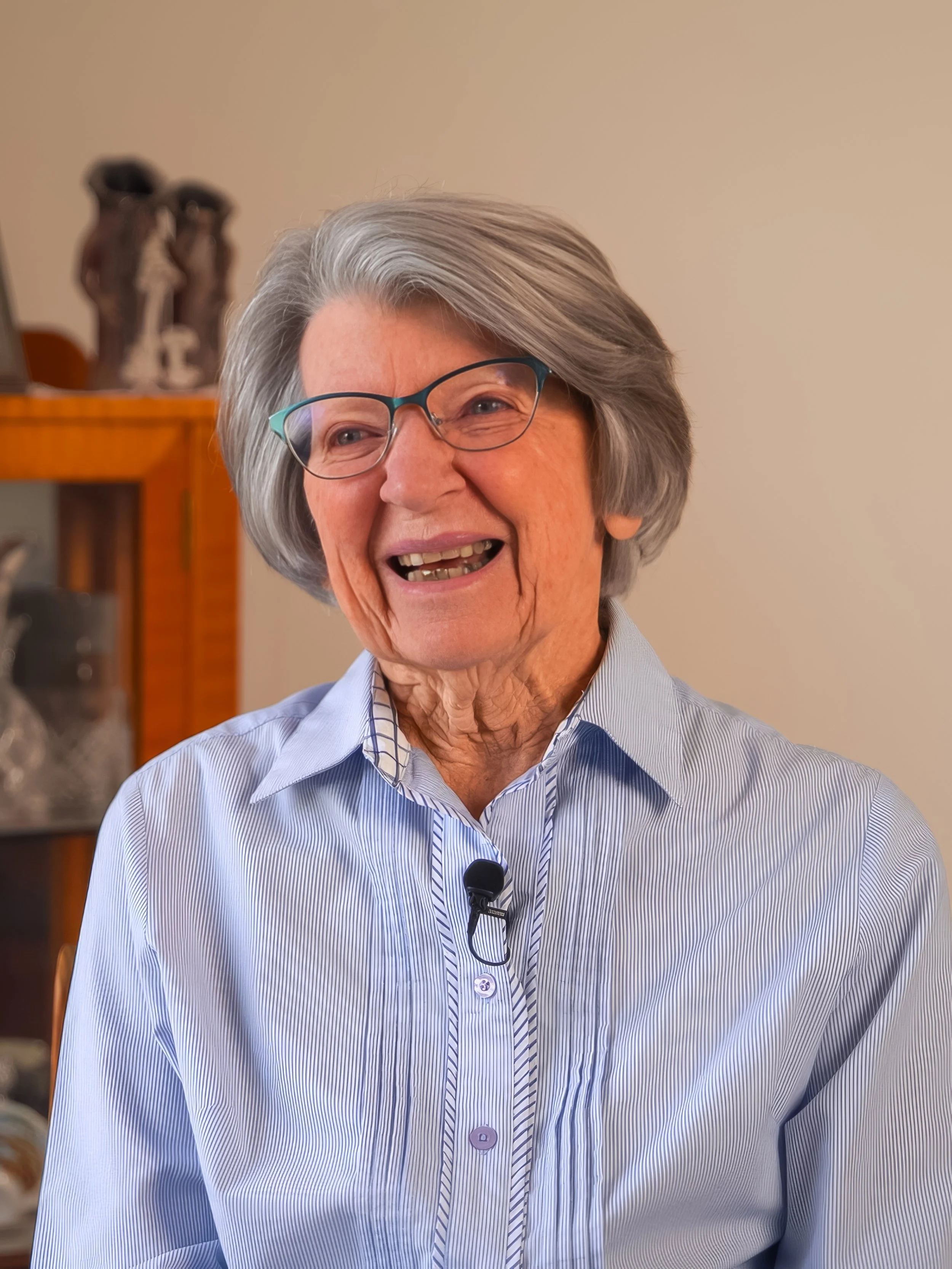 An elderly woman with gray hair and glasses, smiling and wearing a striped blue shirt, sitting indoors.