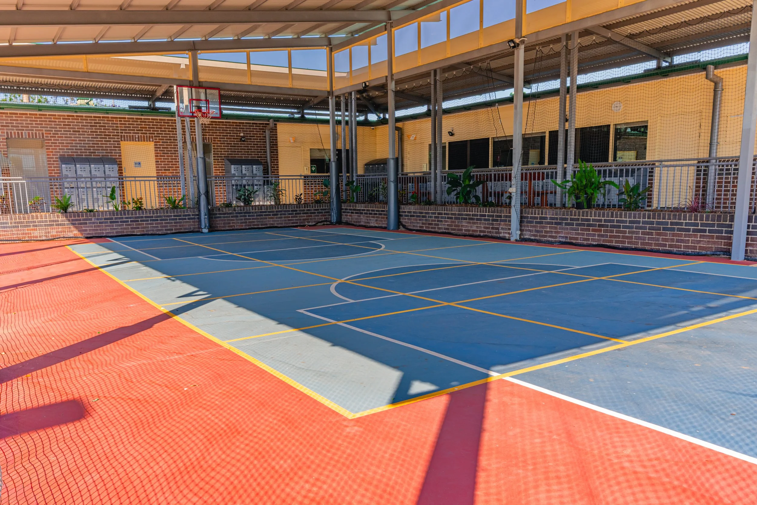 An outdoor basketball court with a blue playing surface and surrounding red border, enclosed by a black fence, with a brick building and apartment balconies in the background, under a yellow metal canopy.