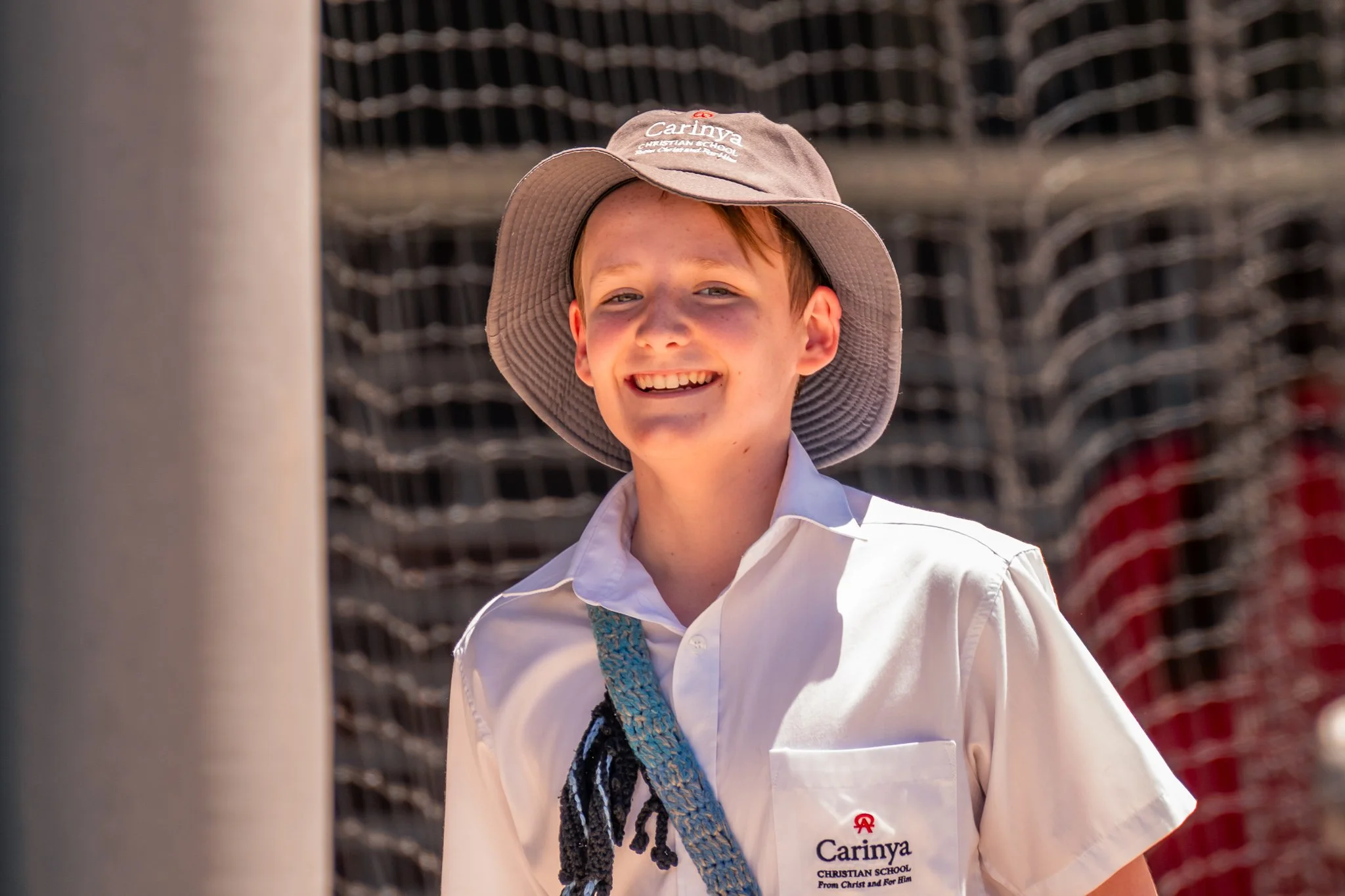 A young boy smiling, wearing a white shirt with a 'Cariyna Christian School' logo, a wide-brimmed hat, and a lanyard, standing outdoors with a wire fence in the background.