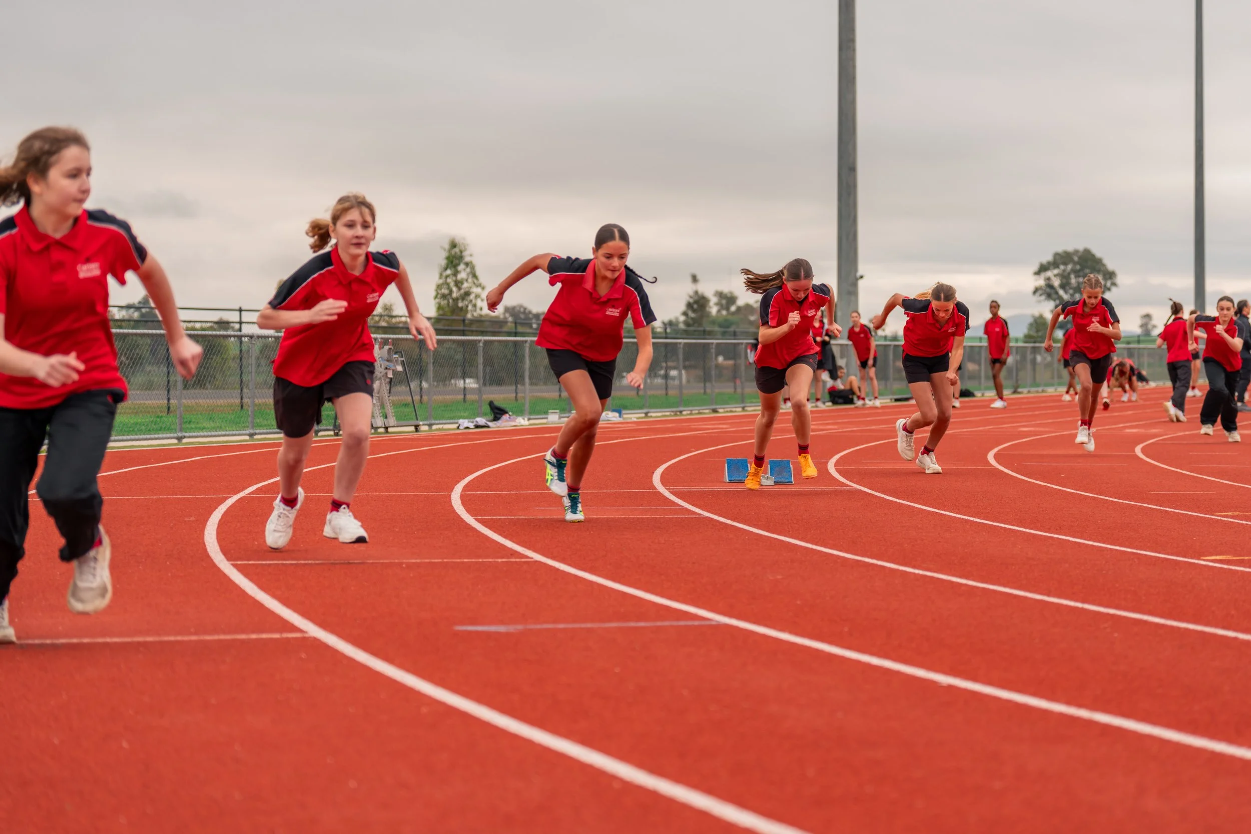 Young girls in red and black sports uniforms sprinting on a red running track during a race.