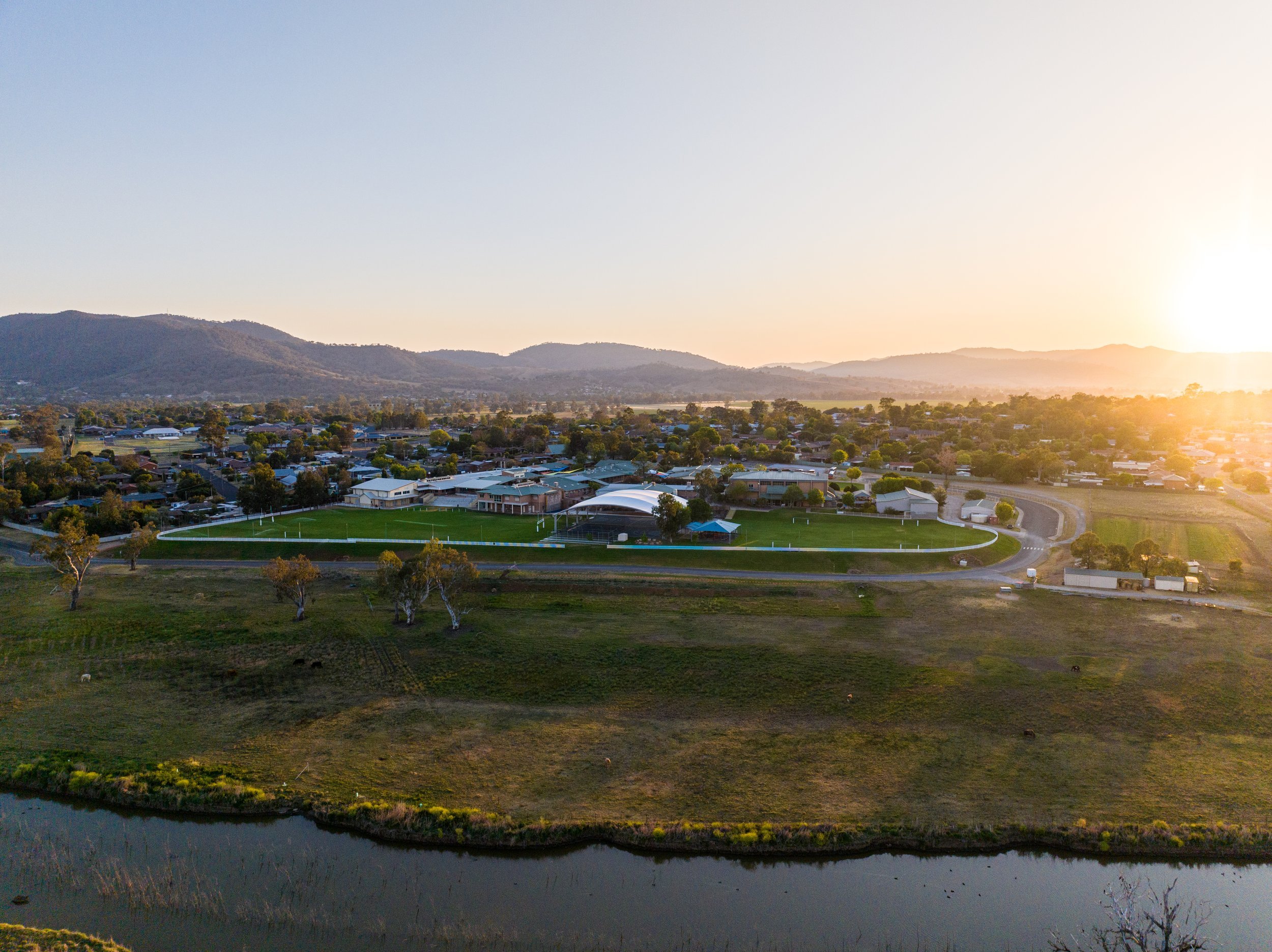 Aerial view of Carinya Christian School in Calala, NSW, showing a sports field and school buildings at sunset, mountains in the distance, and a body of water in the foreground.