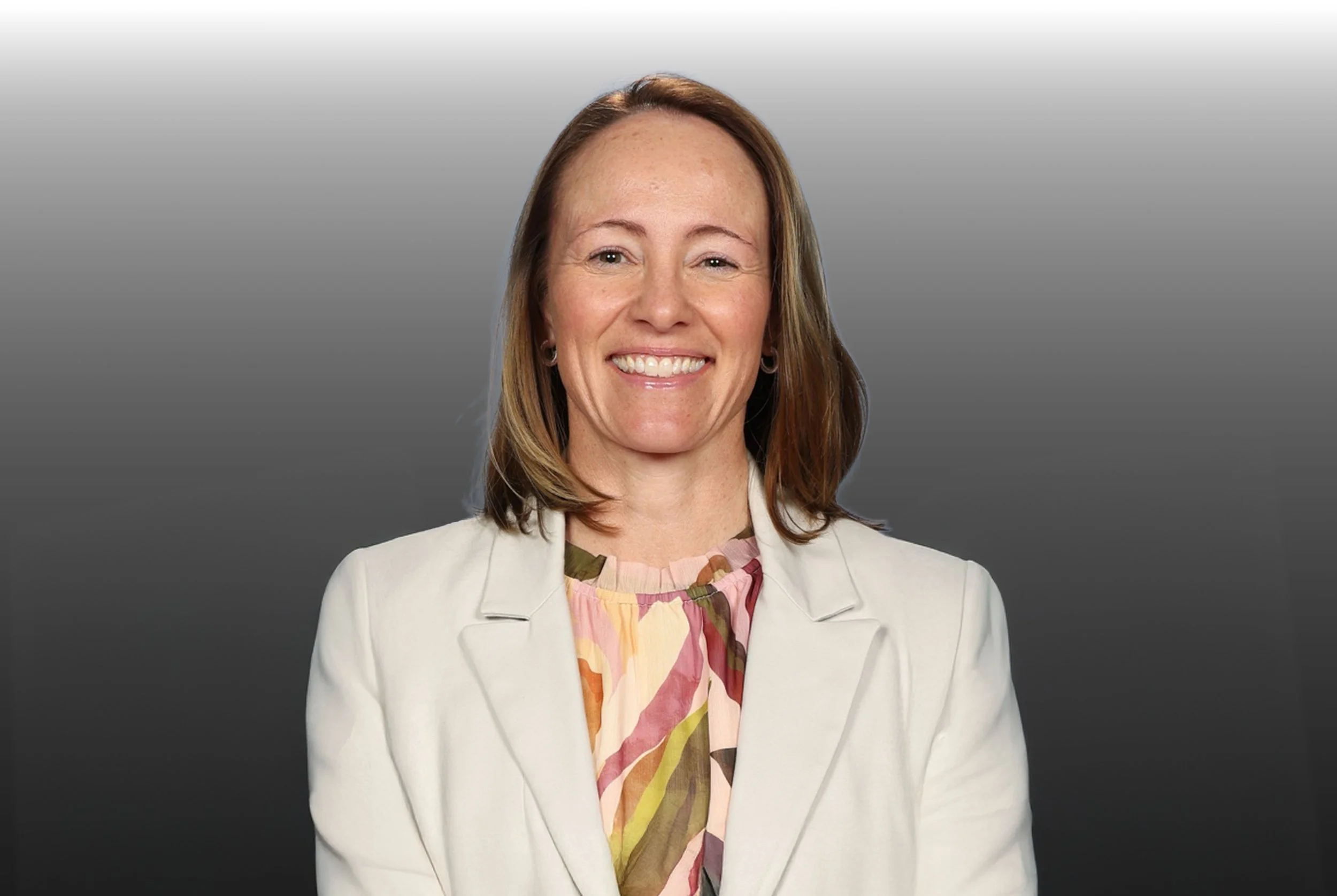 Professional woman with shoulder-length brown hair, wearing a white blazer and a colorful, patterned blouse, smiling against a gray gradient background.