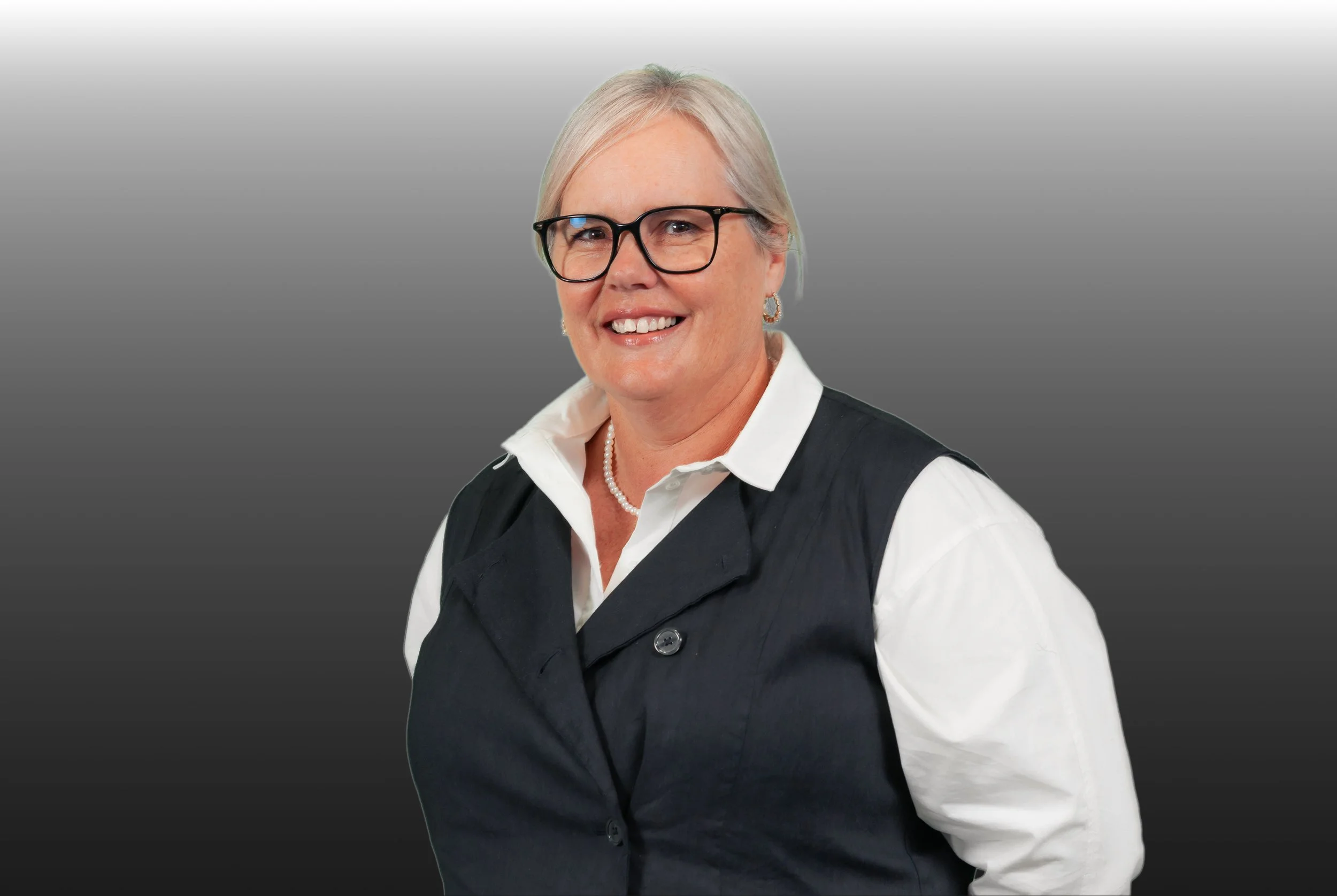 A woman with short white hair and glasses smiling, wearing a white shirt and black vest, against a gray gradient background.