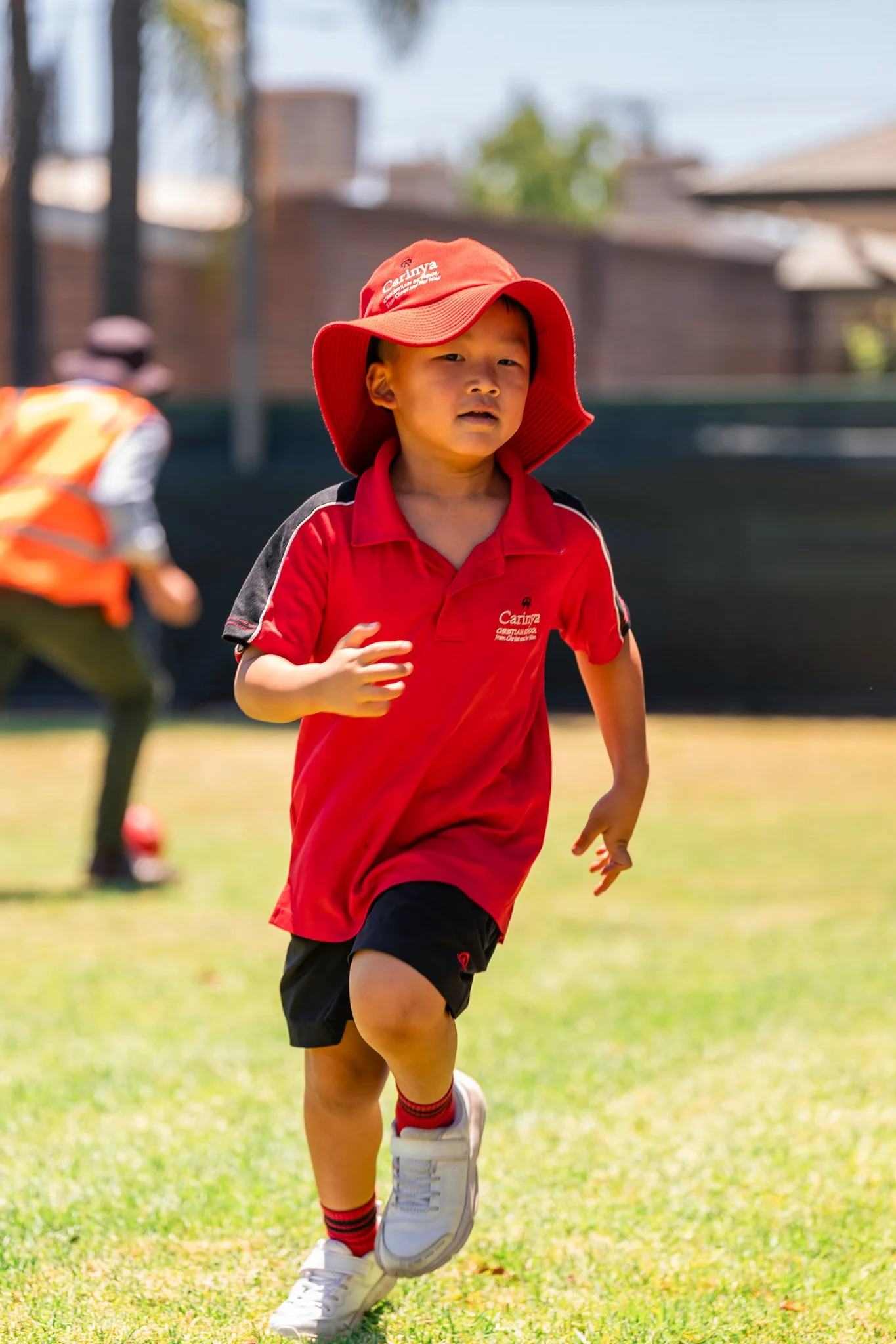 A young boy running on a grassy field, wearing a red shirt, black shorts, white sneakers, and a red hat, with another child in the background.