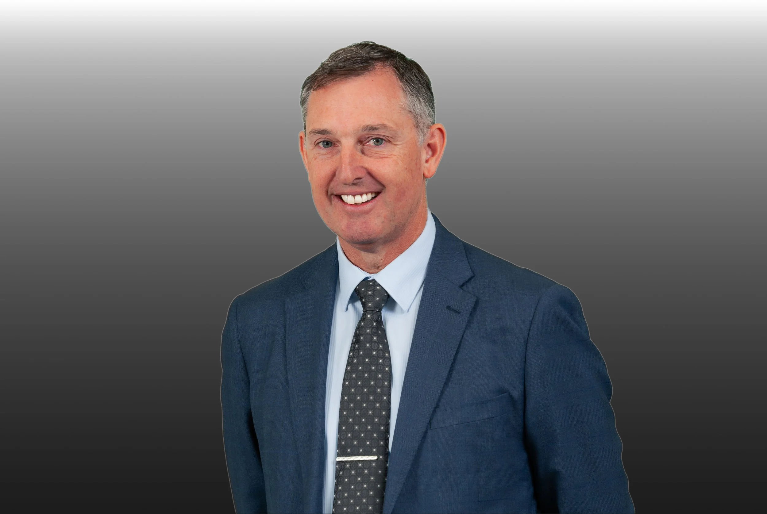 A well-dressed man in a blue suit, white shirt, and patterned tie, smiling, against a gray gradient background.