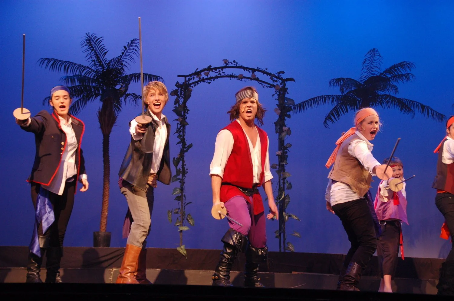 Stage performance of children dressed as pirates with palm trees and tropical archway in background, engaging in a lively scene.
