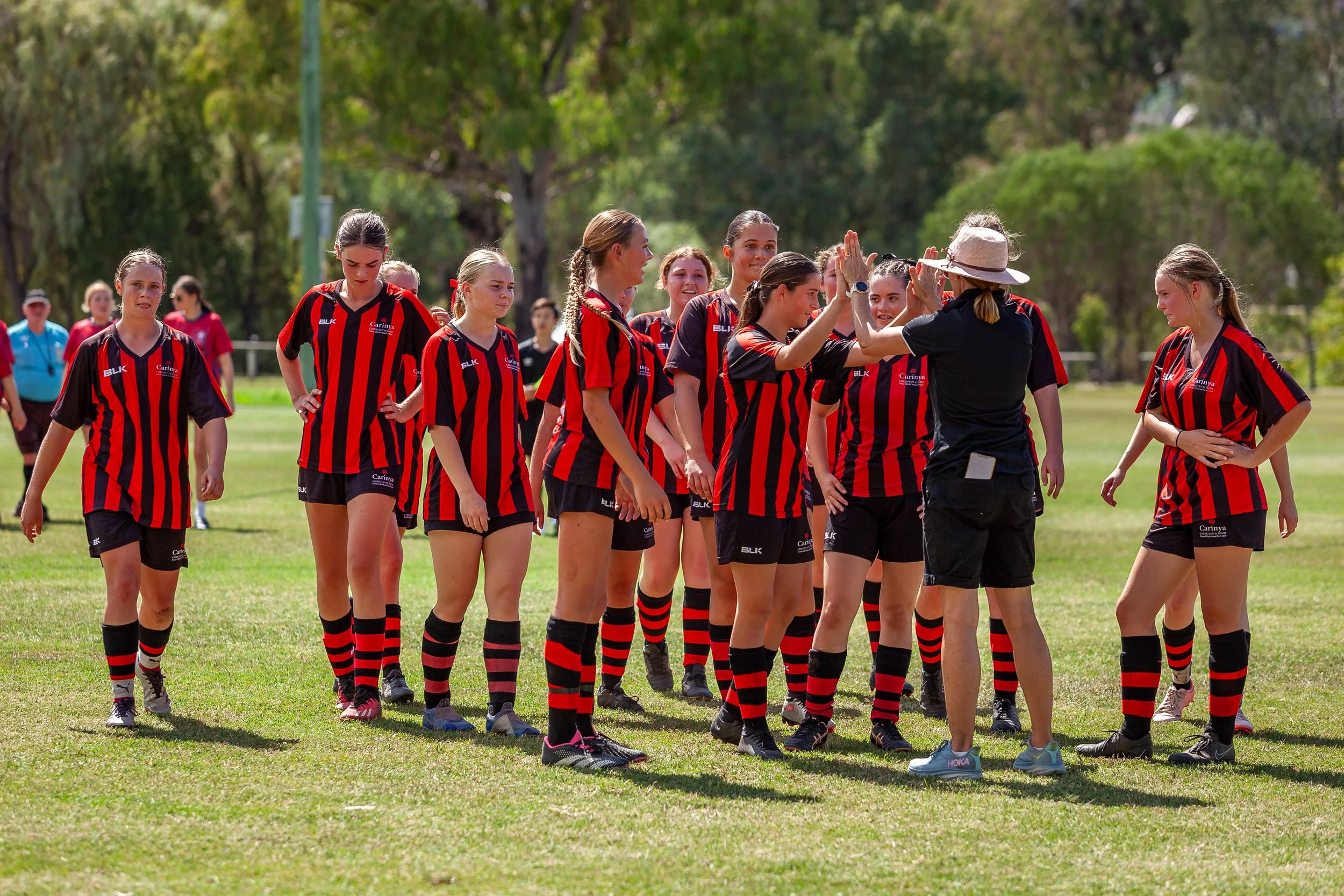 Girls' sports team in red and black striped jerseys high-fiving coach on a grassy field with trees in the background.