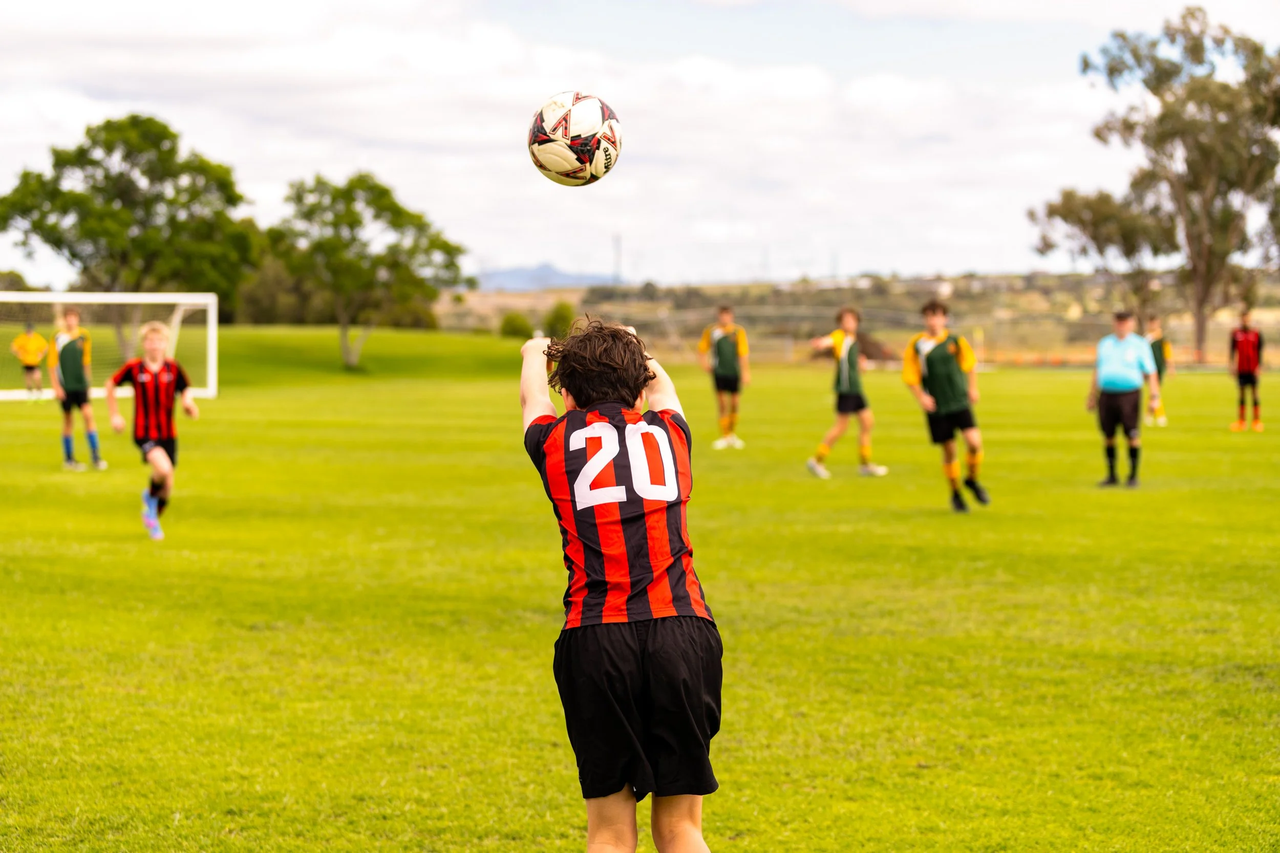 A soccer player with the number 20 on his jersey is taking a free throw during a game on a grassy field, with other players in the background and a soccer goal in the distance.
