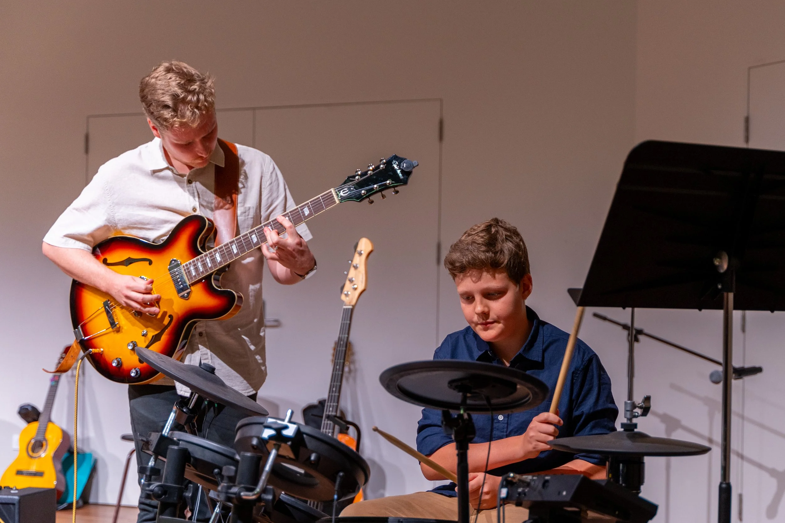 Two young men practicing musical instruments in a room. The one on the left is playing an electric guitar, and the one on the right is sitting at an electronic drum kit, holding drumsticks.