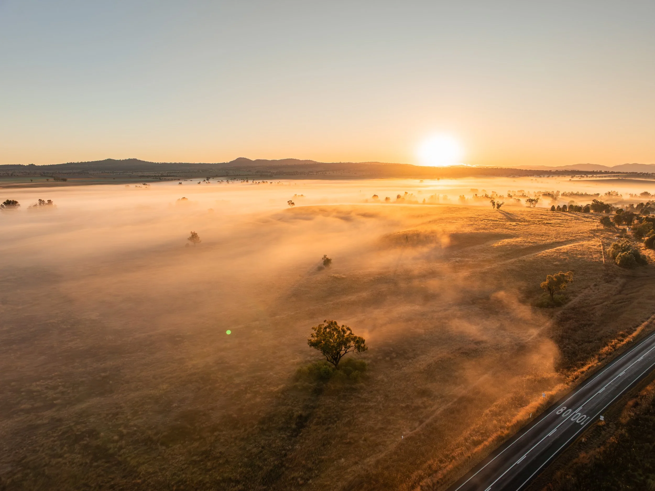 A scenic sunrise over a foggy, rural landscape with scattered trees, a highway in the foreground, and hills in the background.