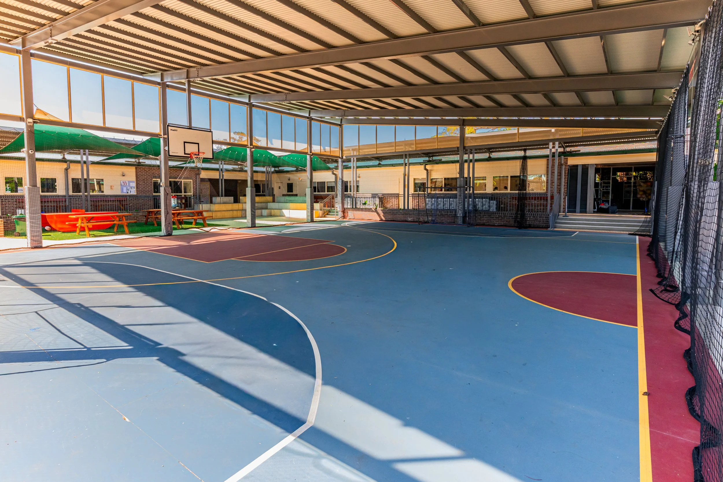 Empty outdoor multi-sport sports court with a basketball hoop, surrounded by fencing and seating, under a partially covered roof on a sunny day.