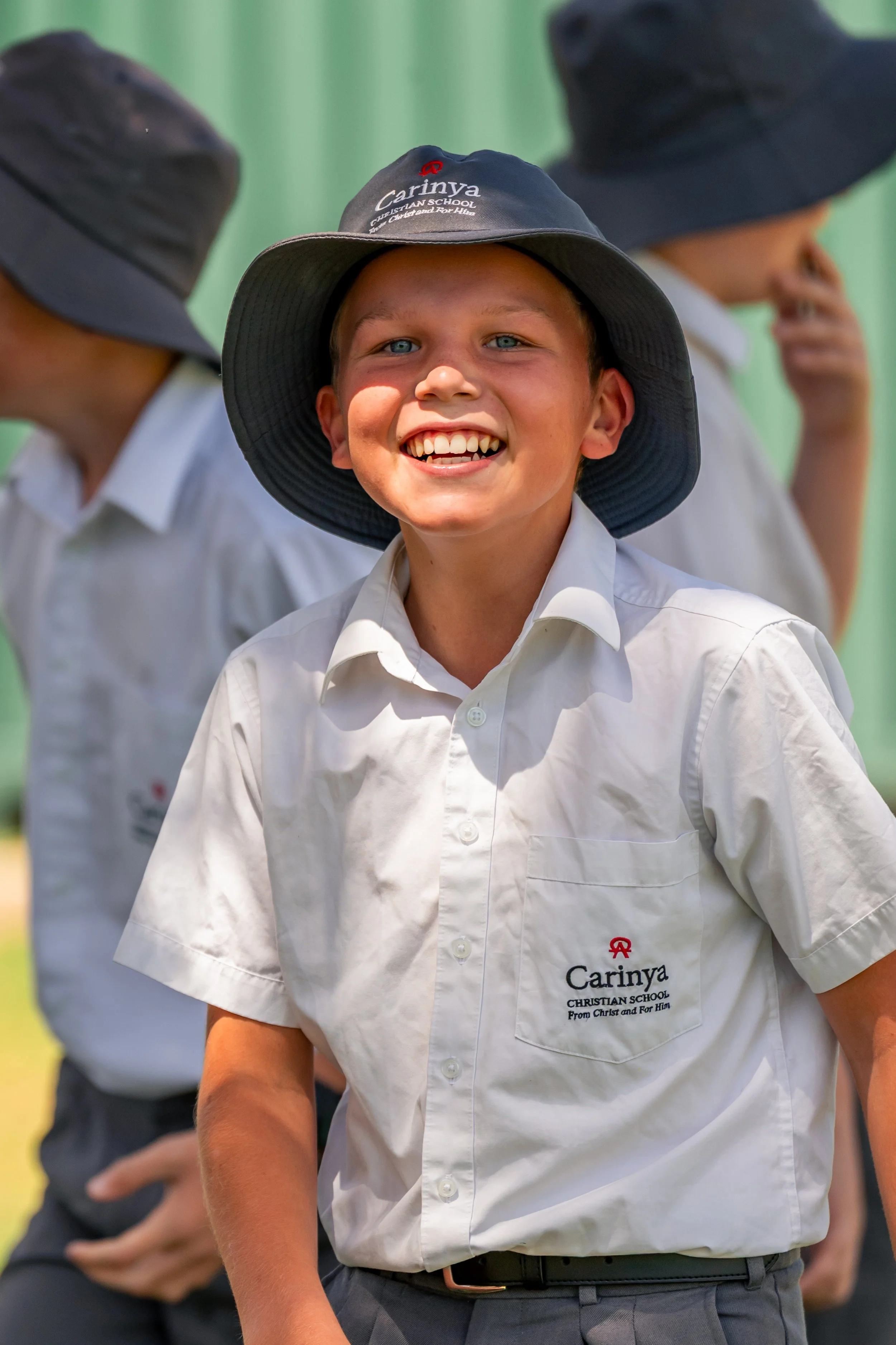 A smiling boy in a white school uniform and gray hat, with other children in similar uniforms and hats in the background.