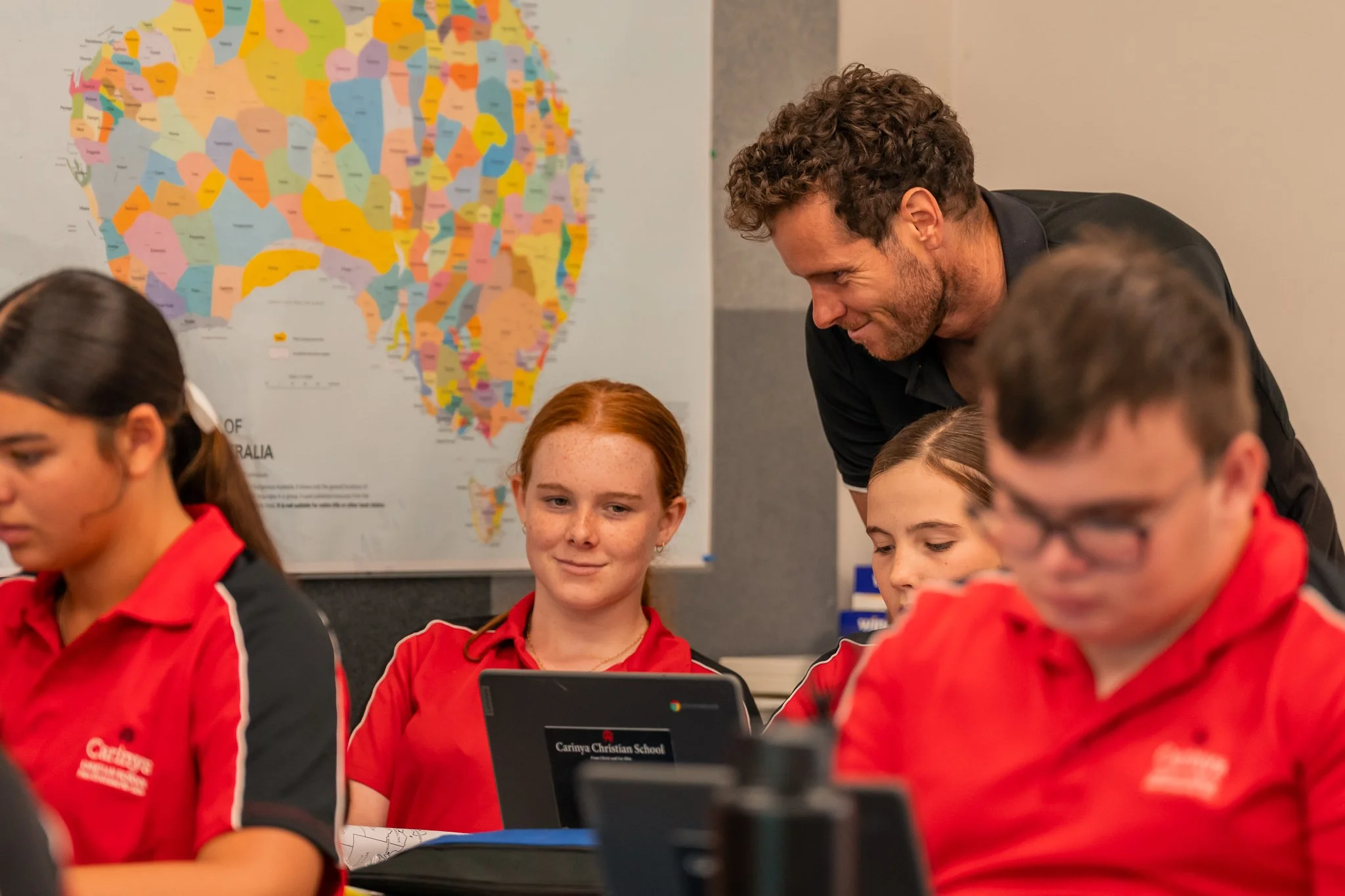 Teacher or instructor assisting students in a classroom with a world map of Australia in the background. The students are wearing red uniforms with school logos, and one student is using a Chromebook.