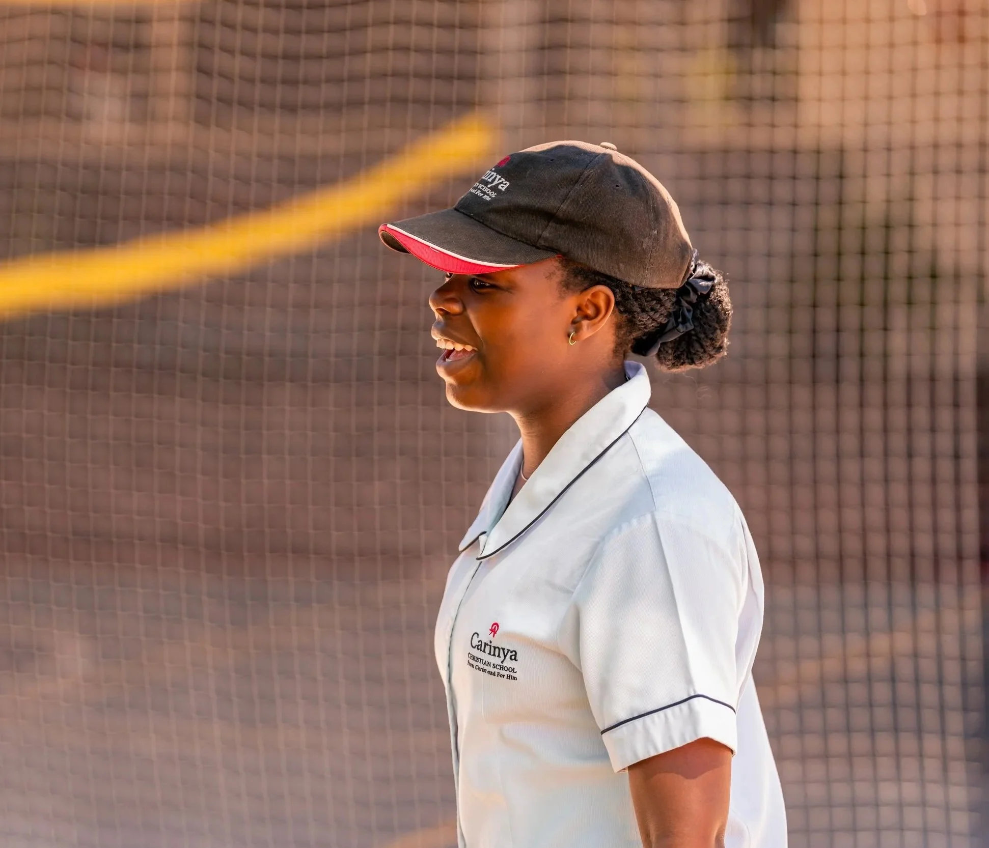 A young woman with dark skin, wearing a white shirt with black trim and a black and red baseball cap, smiling outdoors near a net, possibly at a sports field.