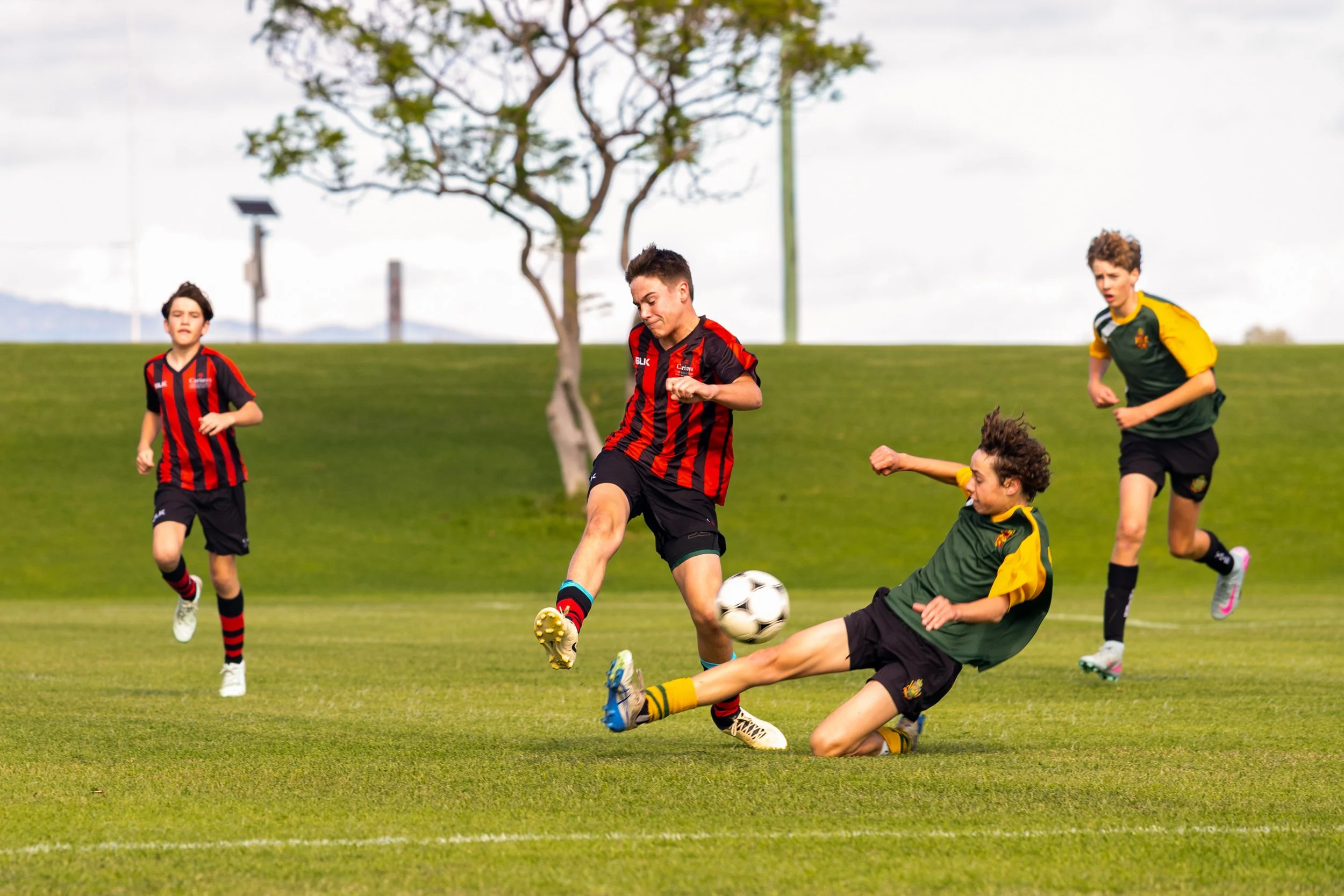 Young boys playing soccer on a grassy field during daytime, with one player in a red and black uniform kicking the ball while a goalkeeper in green and yellow attempts to block a shot, and two other players in the background.