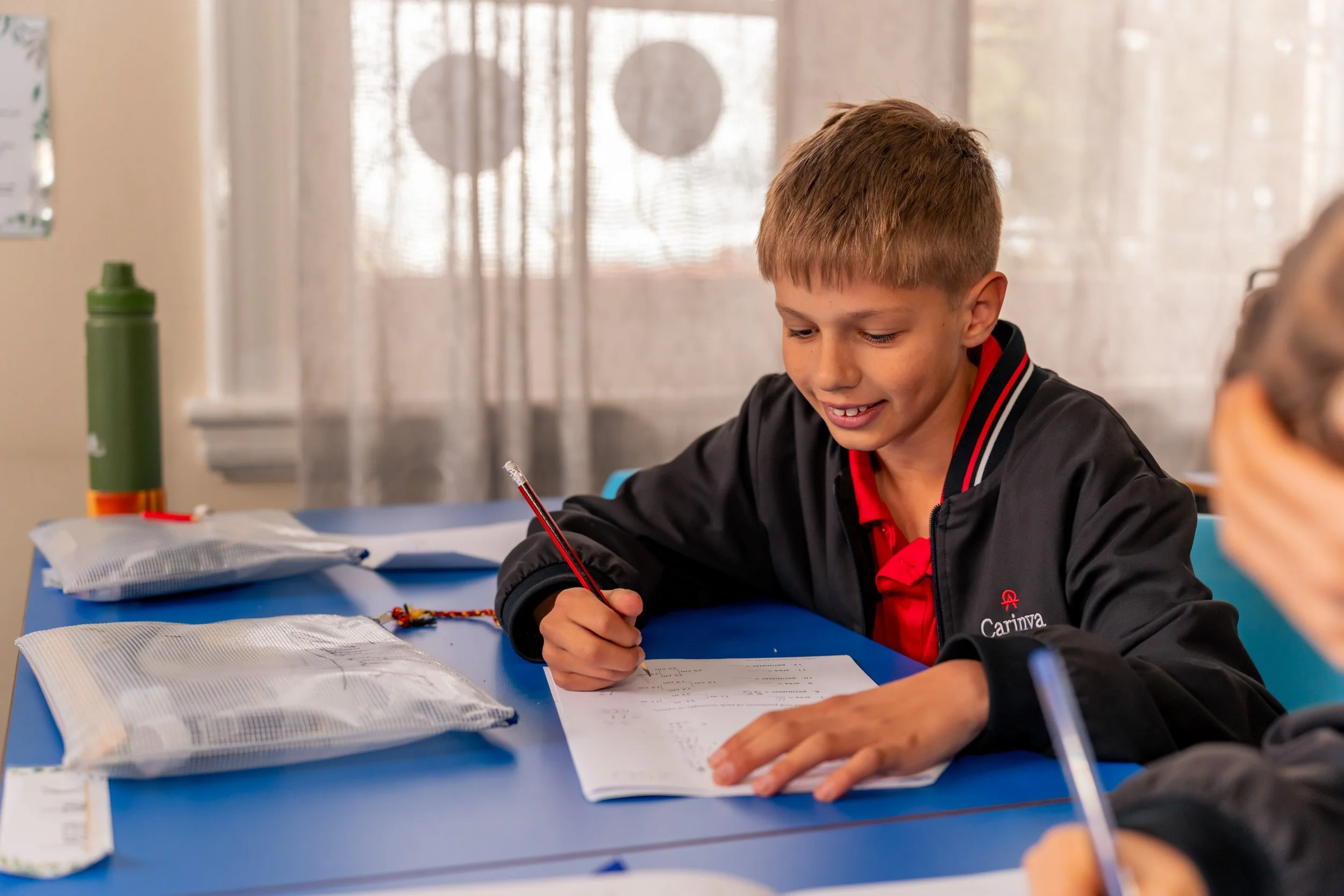 A boy sitting at a blue table, writing in a notebook with a red pen, surrounded by packages and school supplies in a classroom.