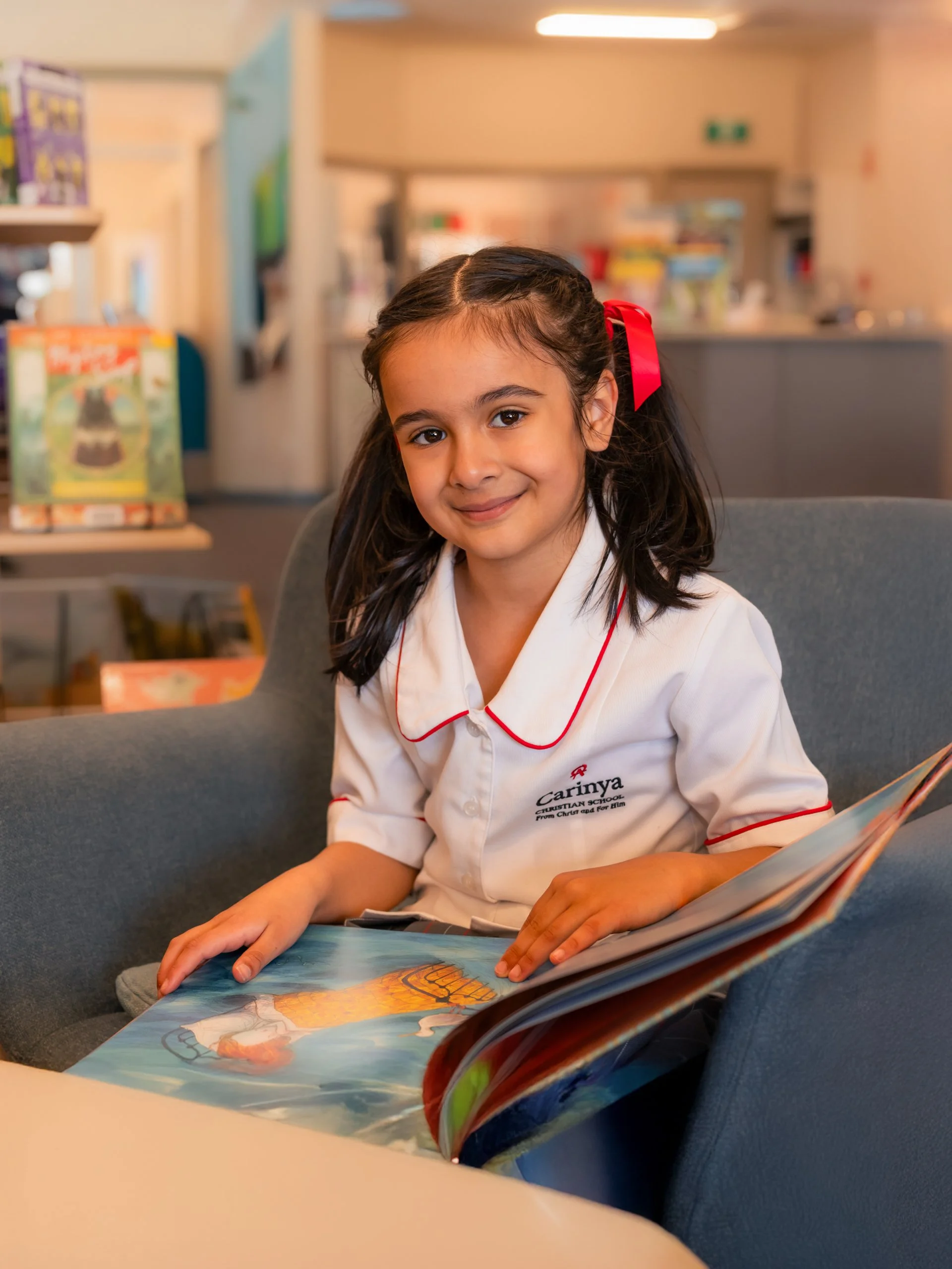 A young girl with dark hair tied back with a red ribbon, wearing a white school uniform with red trim, is sitting on a gray sofa and reading a colorful picture book in a library or classroom setting.