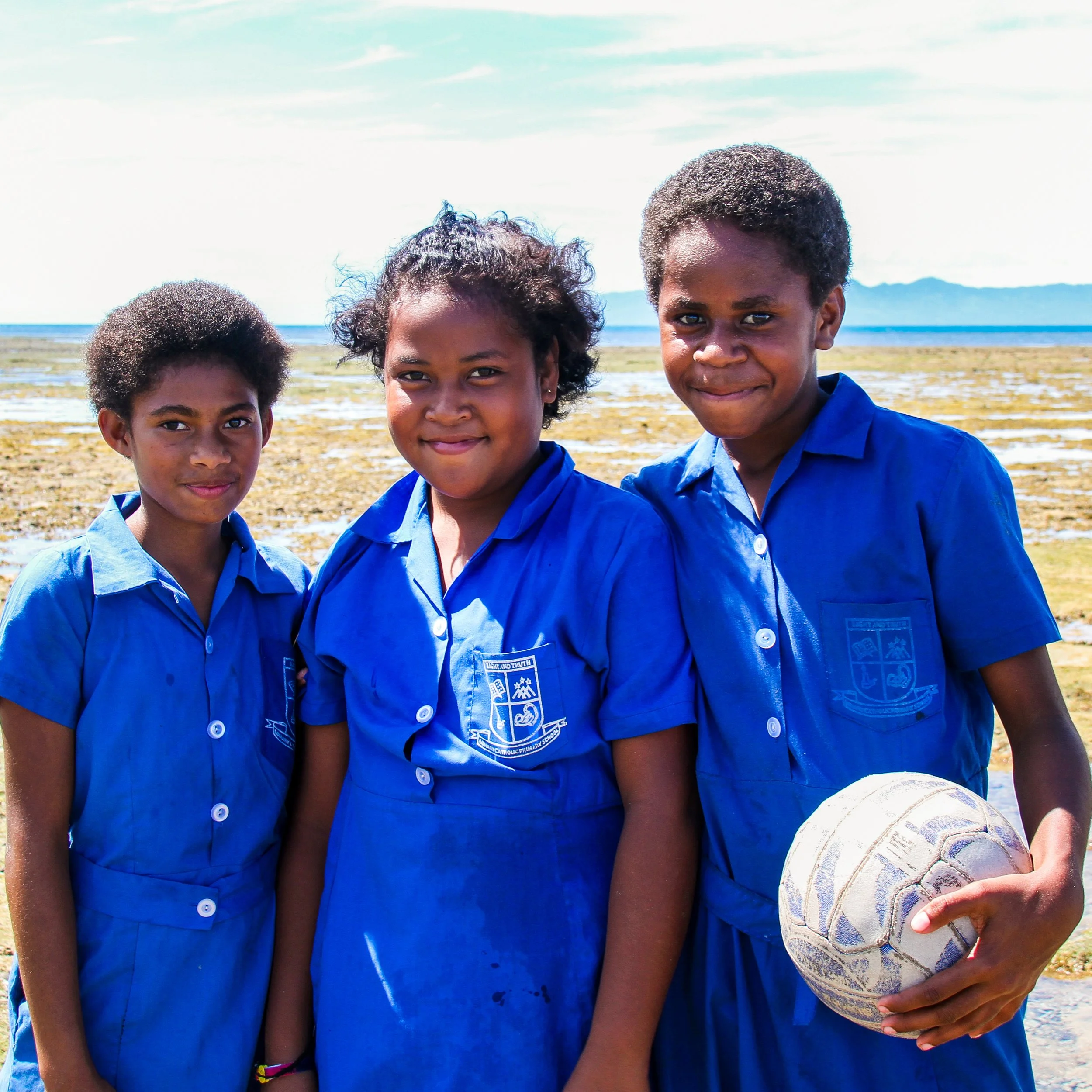Three children in blue school uniforms standing outdoors on a sunny day, with a coastline in the background; one boy is holding a worn volleyball.