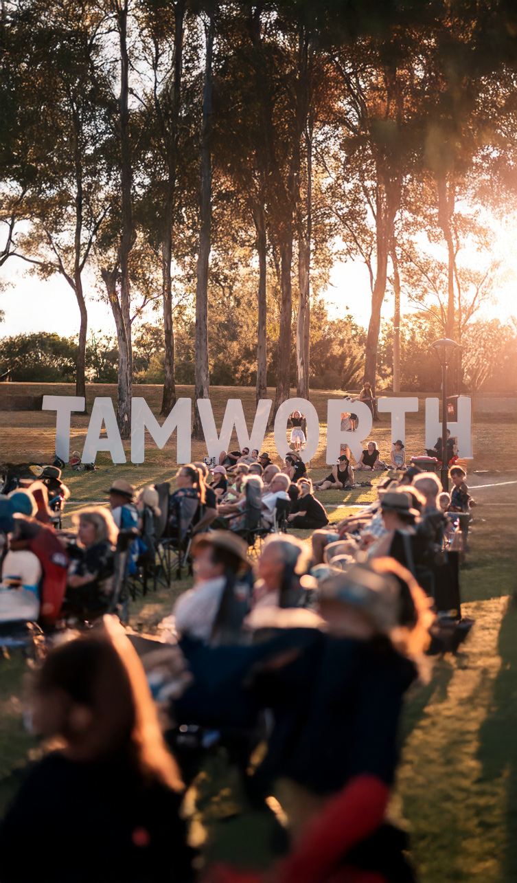 People sitting outdoors on chairs and blankets during sunset, gathering near large white letters spelling 'TAMWORTH' with trees in the background.