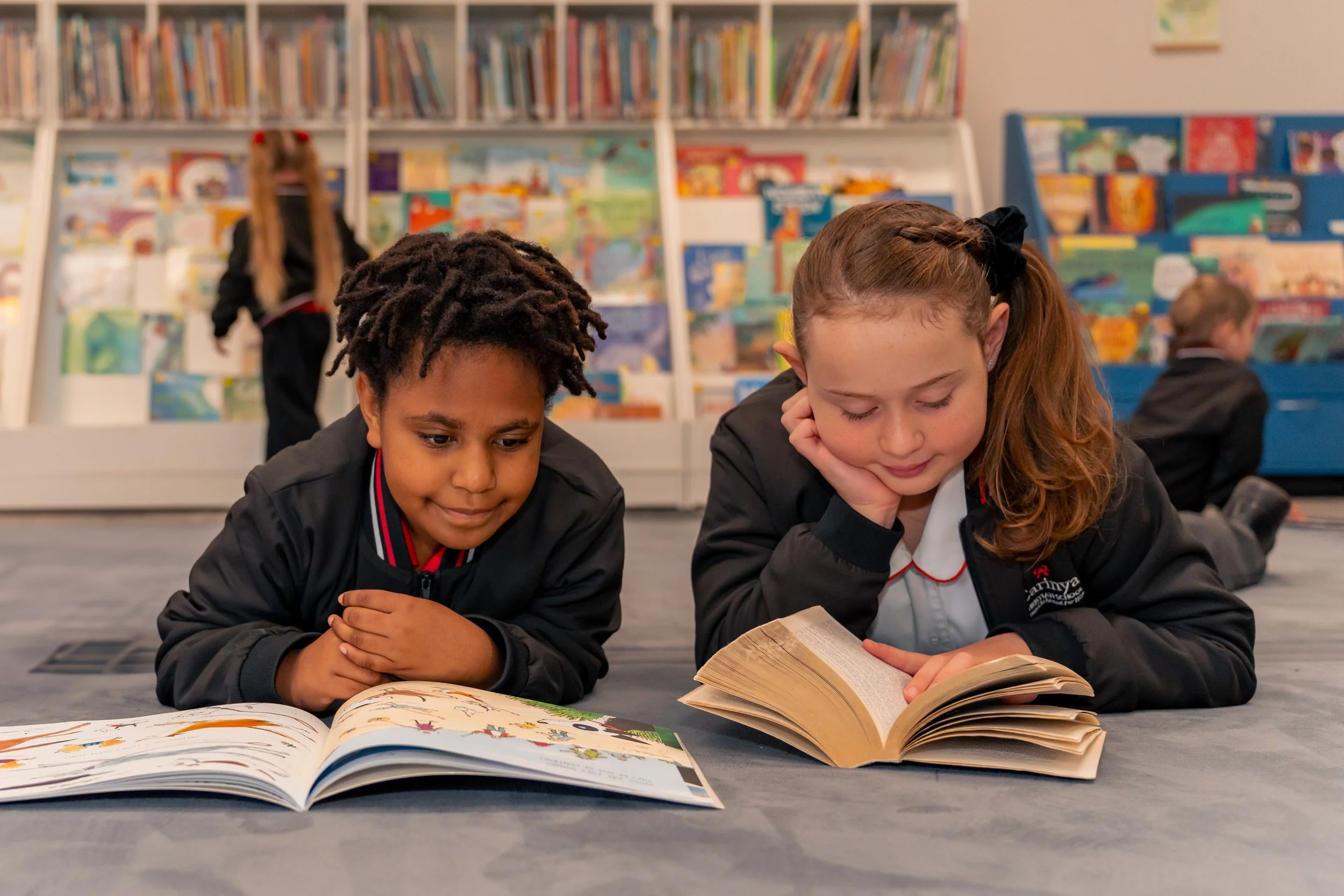 Two children, a boy with curly hair and a girl with a ponytail, lie on the floor reading books in a library or classroom with bookshelves in the background.