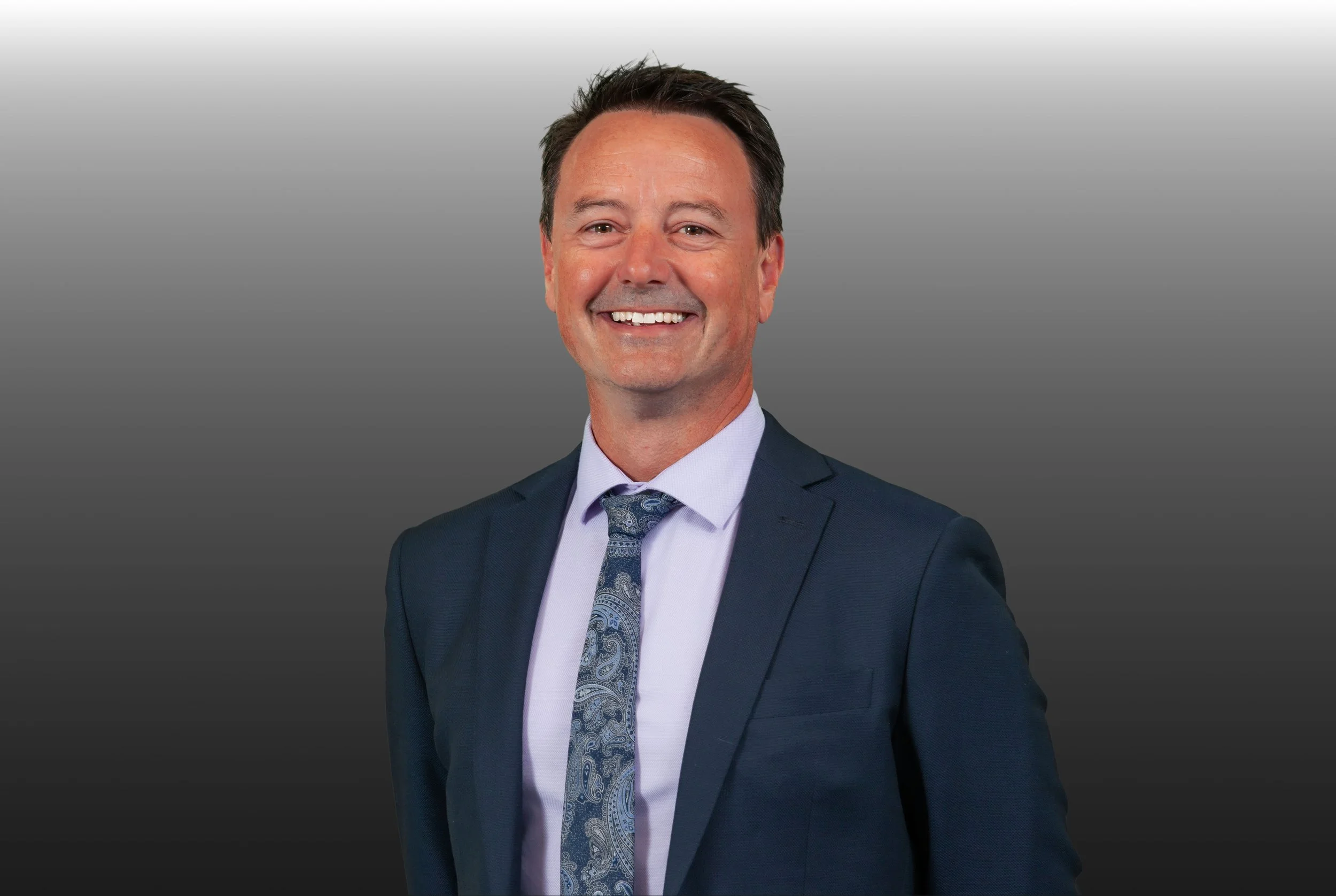 A smiling man in a navy suit and tie standing against a gray gradient background.