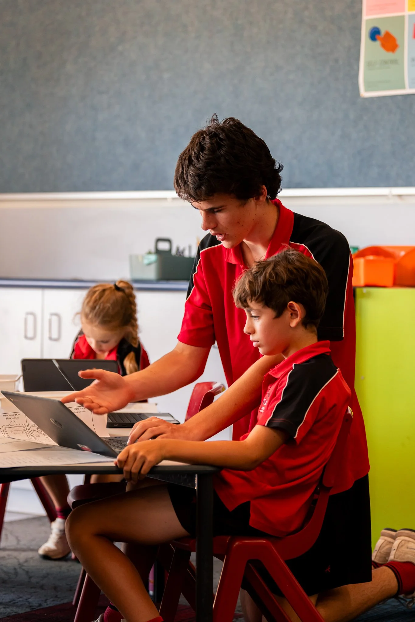 A young male teacher assisting a young student with a laptop in a classroom. Other students are in the background.