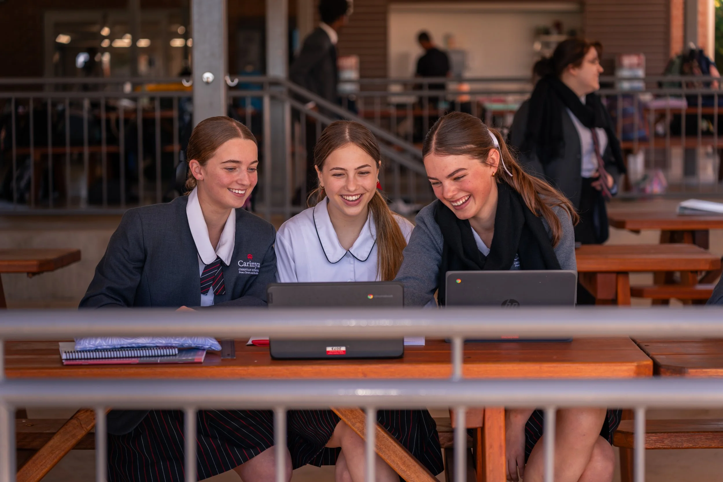 Three smiling schoolgirls in uniforms sitting at a wooden table outside, looking at a laptop and engaging with each other.