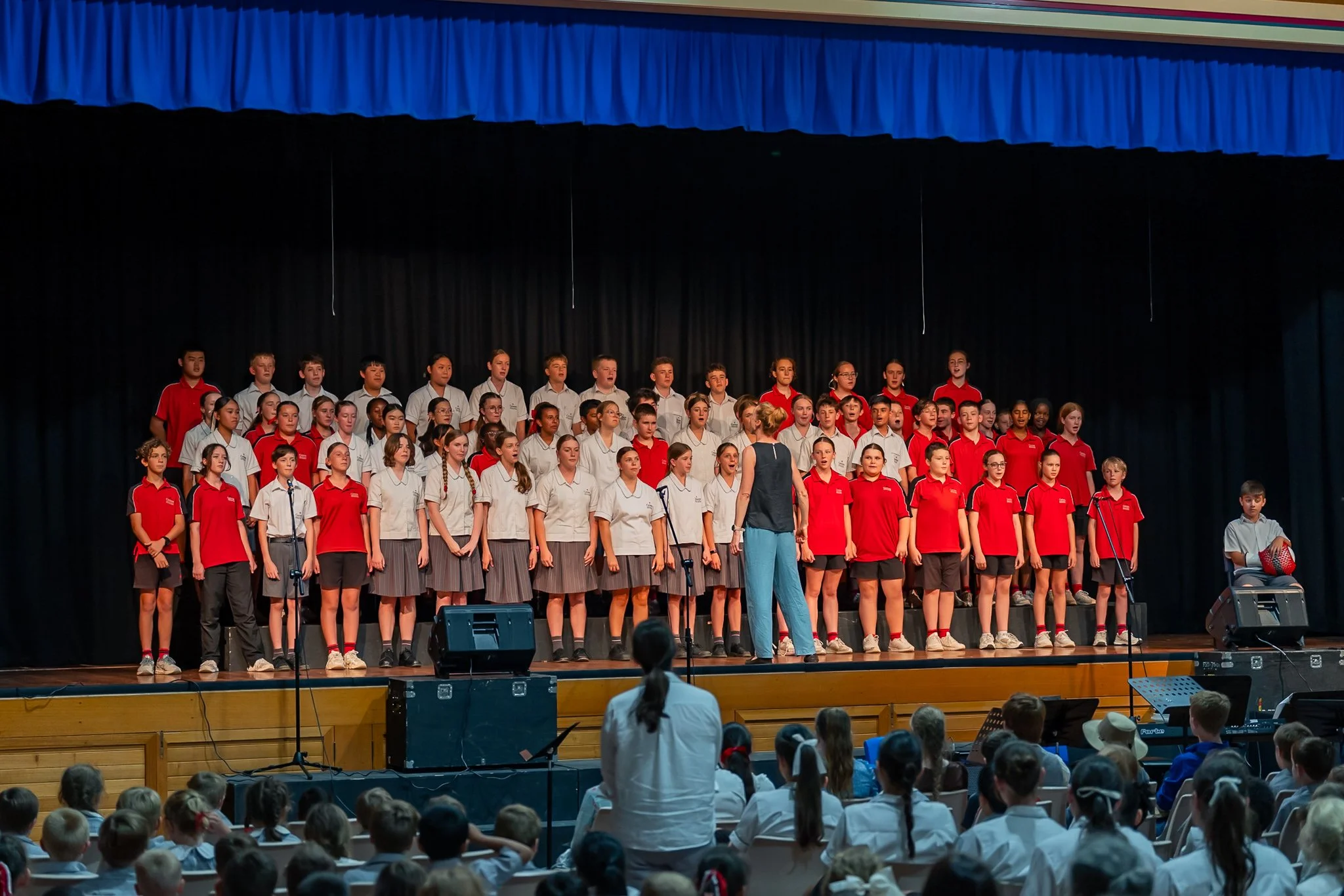 Group of children performing on stage with a conductor facing them, audience watching, dark curtain backdrop.