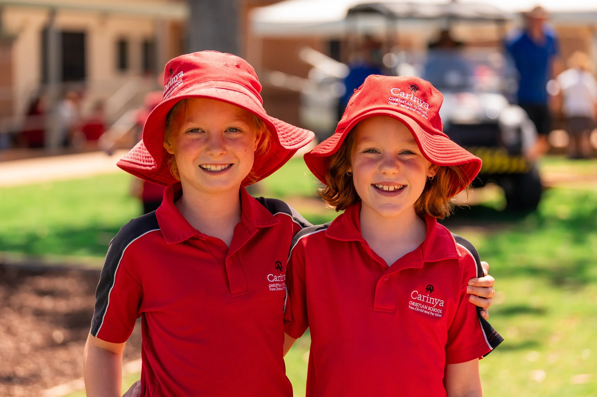 Two young girls wearing red hats and red polo shirts with 'Carinya Christian School' logo, smiling outdoors on a sunny day.