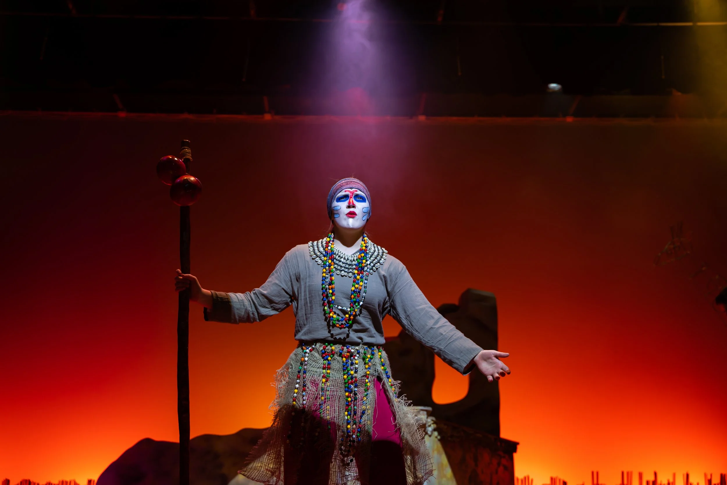 Performer with painted face, colorful bead necklaces, and traditional costume, holding staff with ornaments, on stage with red-orange background.