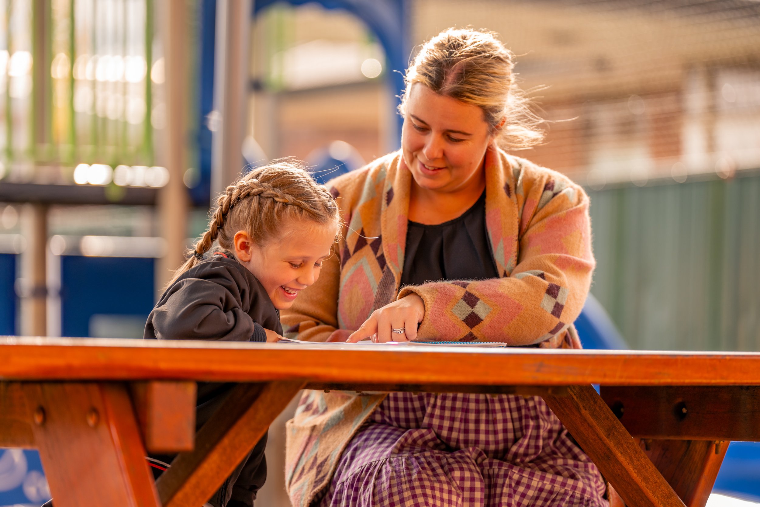 A woman and a young girl sitting together at a wooden table outdoors, smiling and looking at something on the table.