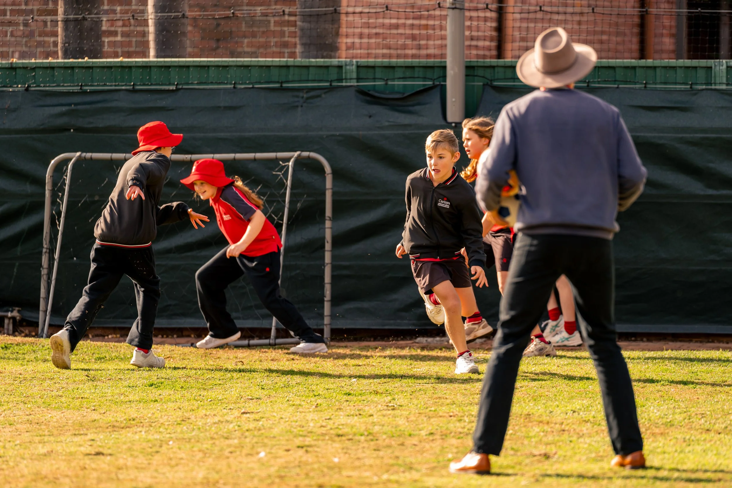 Kids playing soccer on a field, with one child kicking and others running, while a person with a hat watches.
