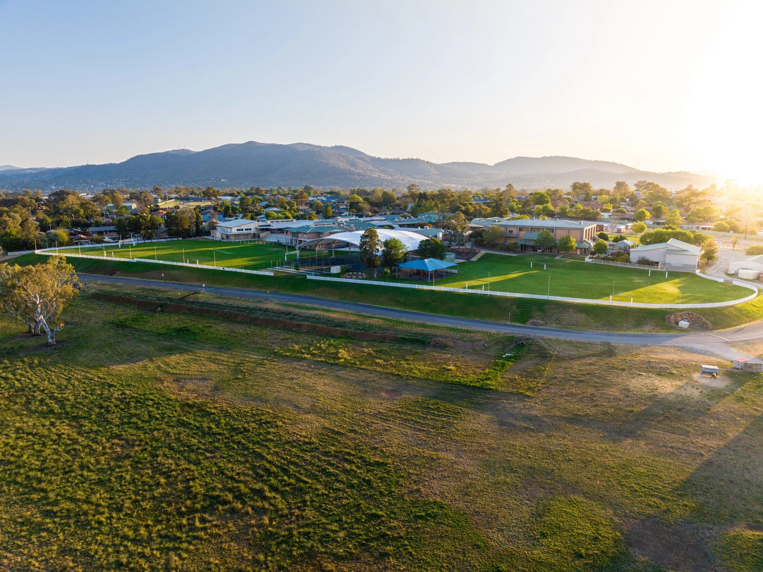 Aerial view of a school with a large sports field, surrounded by trees and residential buildings, with mountains in the background and the sun setting on the horizon.