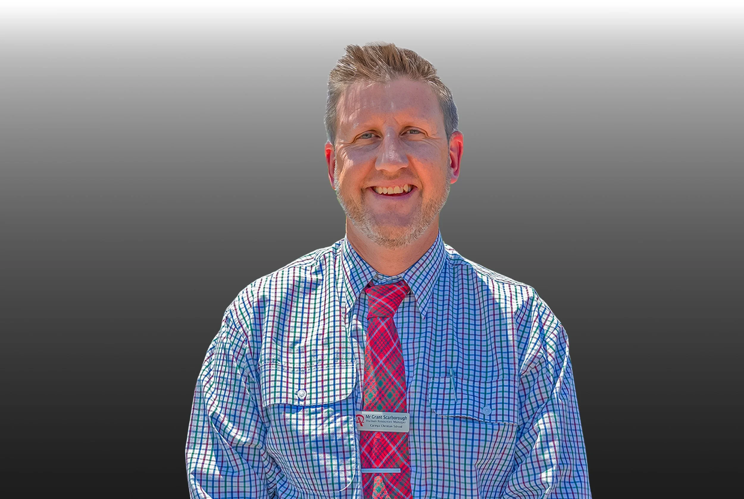 A man with short blond hair, a beard, and mustache smiling, wearing a checkered shirt and a red plaid tie, standing outdoors against a gradient gray background.