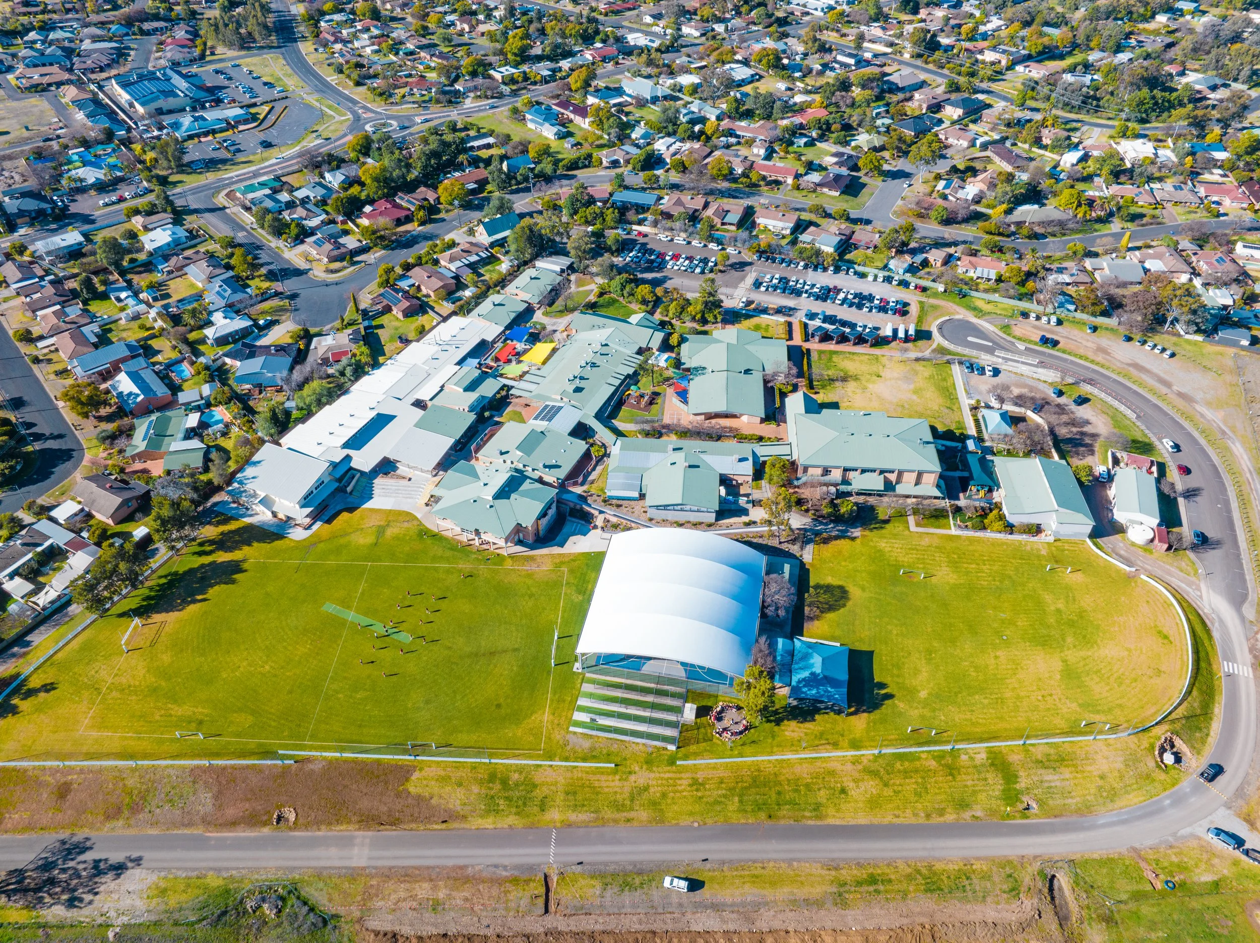 Aerial view of Carinya Christian School in Calala, NSW, showing a sports field and school buildings at sunset, mountains in the distance, and a body of water in the foreground.