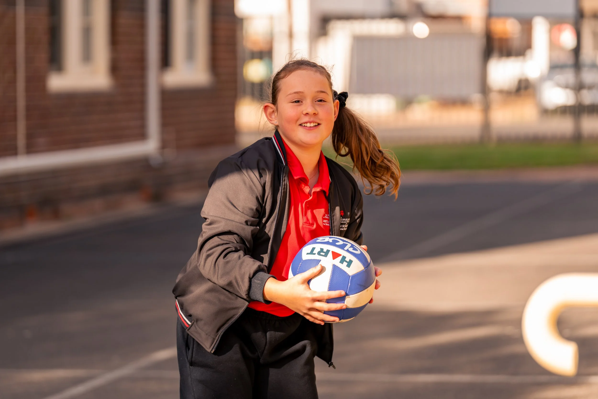 Young girl with a ponytail holding a volleyball on an outdoor court, smiling at the camera