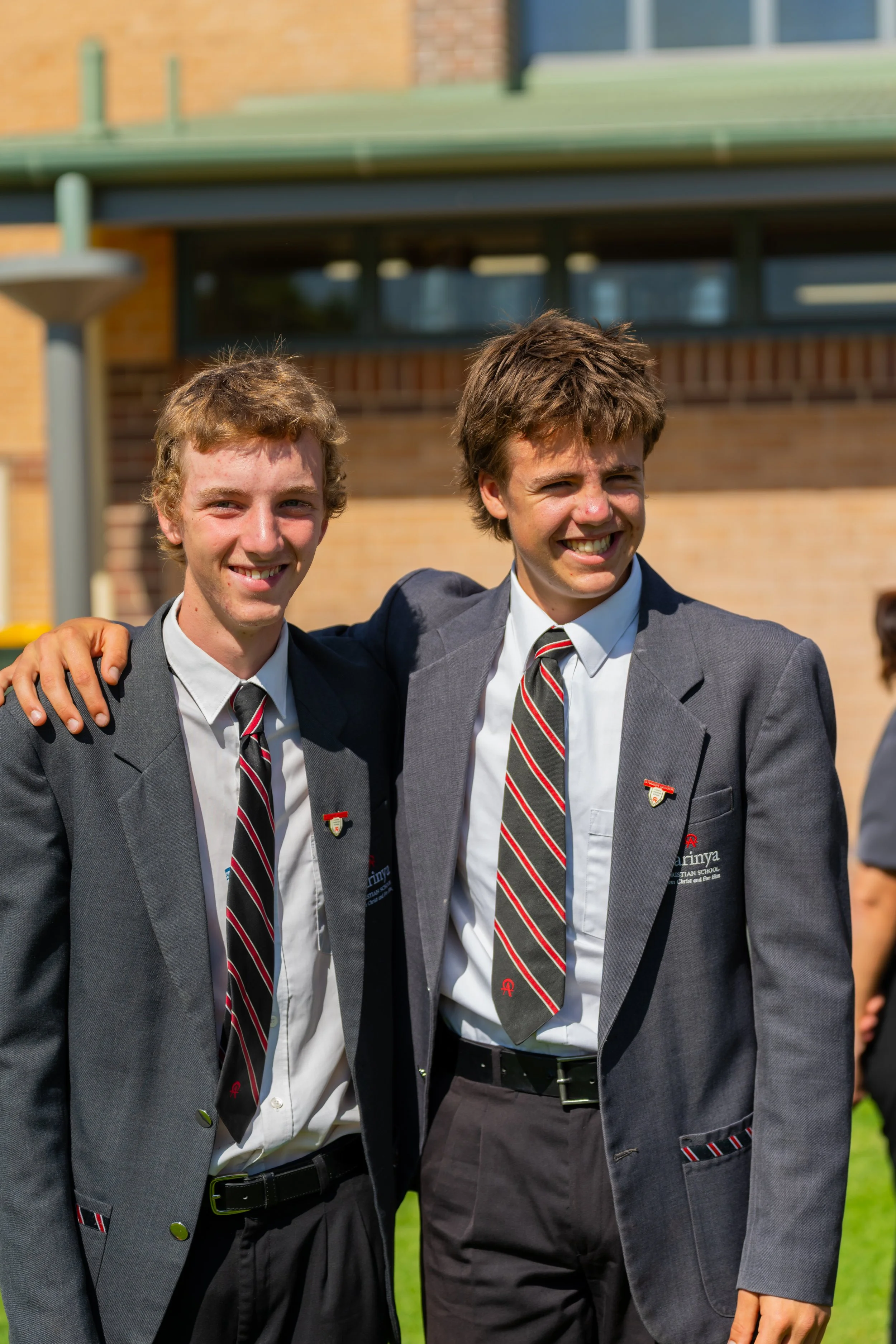 Two teenage boys in school uniforms standing outside with their arms around each other, smiling.